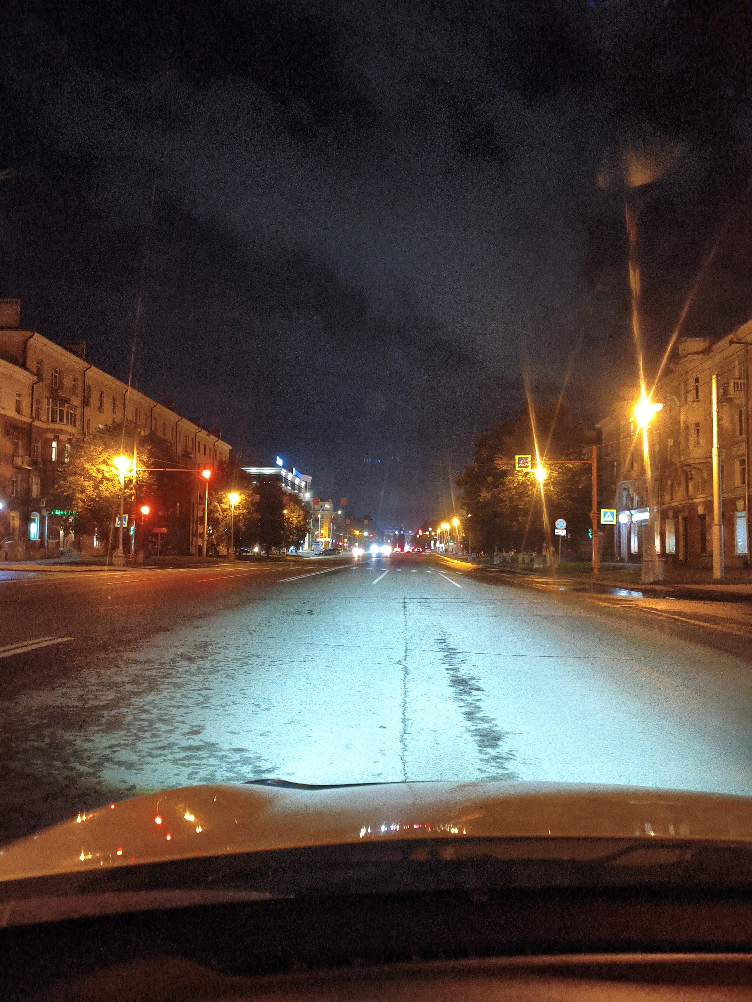 The image depicts a nighttime view of a city street in Kemerovo, Russia, as seen from the interior of a vehicle. The street is wet, reflecting the bright orange glow of streetlights and the headlights of other vehicles. The sky is dark and cloudy. On either side of the street are multi-story buildings, some with illuminated windows. Traffic lights are visible, with one displaying a red signal. Trees line the sidewalk. The foreground shows the dashboard and windshield of the vehicle, with reflections of lights.