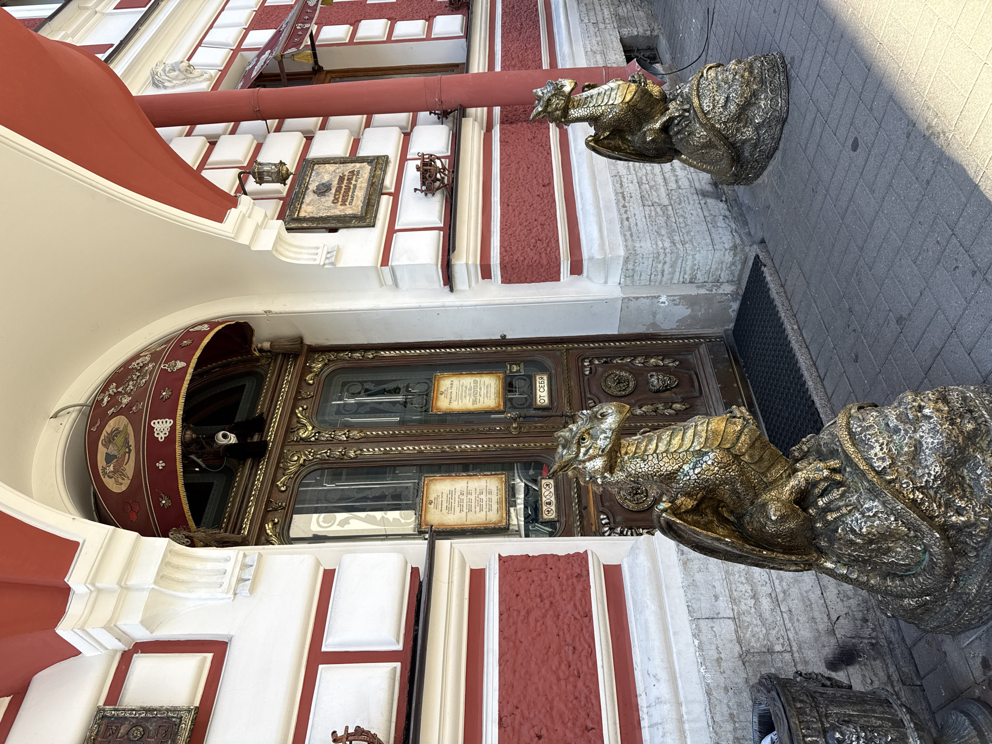 The image shows a view of an ornate building entrance in Saint Petersburg, Russia. Two large, golden dragon statues guard the entrance. The building's facade is painted in alternating bands of white and a deep red, with decorative white molding. Above the entrance, a red awning with gold trim extends over a segment of the facade. The entrance itself features a grand, dark wooden door with intricate carvings and multiple glass panes. Above the door, framed signs with text are visible. The ground in front of the entrance is paved with grey bricks. The lighting suggests it is daytime, with shadows cast by the statues and building. The overall impression is one of historical grandeur and possibly a museum or cultural site.