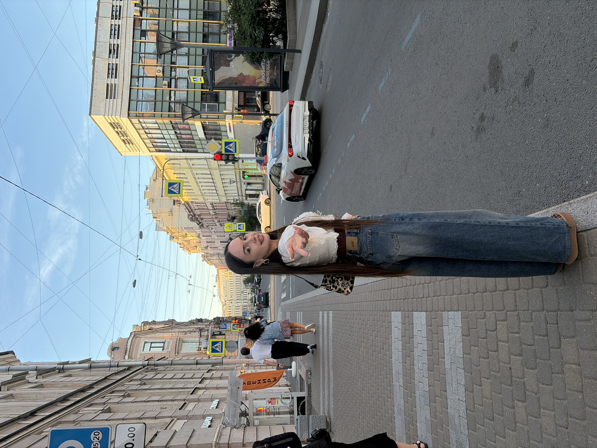 A young woman with long dark hair, wearing a white cropped top and wide-leg blue jeans, stands on a pedestrian sidewalk, turned towards the camera with her right hand extended. Behind her, a street scene unfolds in Saint Petersburg, Russia. The street is lined with buildings of varying architectural styles, some with historical facades and others more modern. Traffic is visible on the road, including a white taxi with red branding and other cars. Pedestrian crossings are marked on the asphalt. Several traffic signs and streetlights are present. The sky above is clear and blue with a few wispy clouds. The lighting suggests it is late afternoon or early evening. A couple walks away from the camera further down the sidewalk.