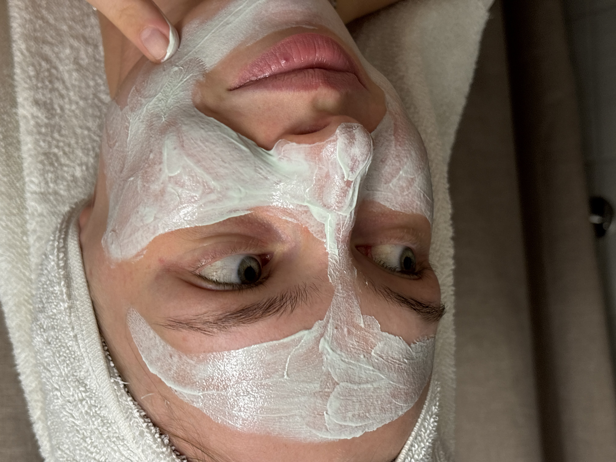 The image presents a close-up view of a person's face undergoing a skincare treatment, likely in Perm, Russia. The individual is lying down, with a white, terry-cloth towel wrapped around their head, securing their hair away from their face. A thick, light-colored, possibly white or pale green, facial mask is applied liberally across their face, covering areas like the forehead, cheeks, nose, and chin. The mask application spares the eye and mouth regions.
The person's eyes are open, with their gaze directed towards the upper right side of the frame, indicating a neutral or slightly observant expression. Their lips are naturally pink and slightly parted. A hand with a neatly manicured finger, featuring a natural or French-tip style nail, is visible near the person's left temple, suggesting an interaction with the mask, perhaps during application or adjustment. The skin visible around the eyes and mouth appears clear and smooth. The background is softly blurred and indistinct, featuring dark, muted tones that hint at an indoor setting, possibly a spa or a personal care space. There is no visible text in the image.