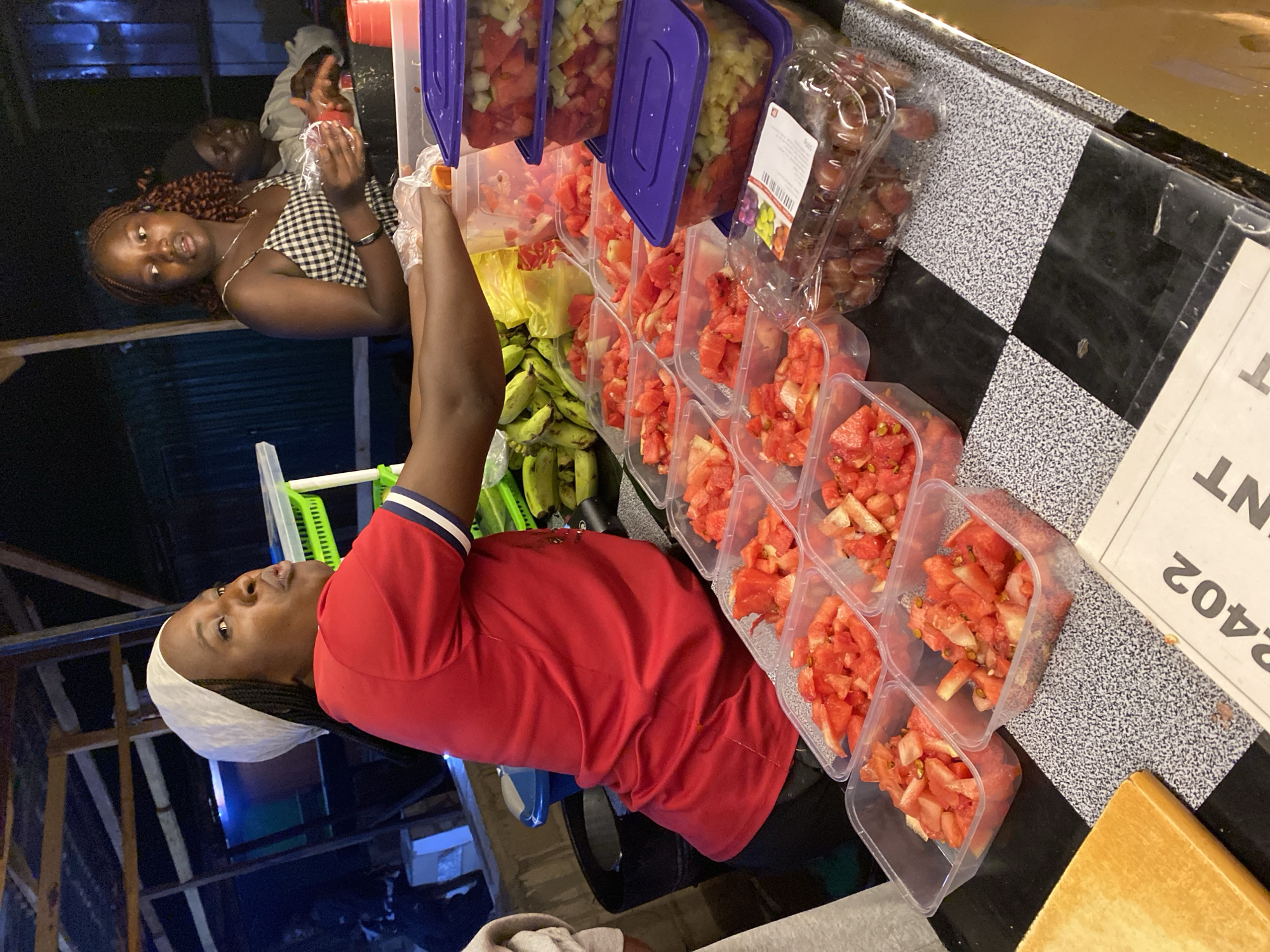 The scene depicts a woman in a red shirt and a white head covering, likely preparing fruit servings at a food stall in Durimi, Nigeria. Several clear plastic containers filled with cut watermelon and other fruits (possibly pineapple) are arranged in rows on a checkered black and white surface. A row of bananas sits behind the containers. There are also larger containers, some with purple lids, stacked with fruit. A woman with braided hair is visible in the background to the left.