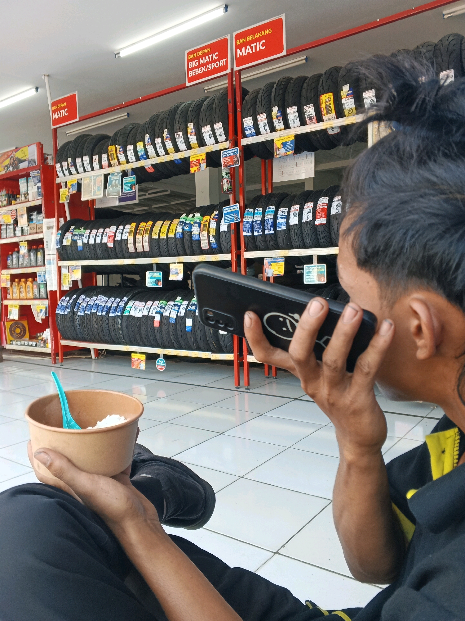 The image captures a man sitting and holding a bowl of rice with a blue spoon, while also holding a black smartphone to his ear, appearing to be on a call. He is wearing a black shirt with yellow accents. The background reveals a well-lit store with shelves stacked high with motorcycle tires. Red signage is visible above the tire displays, indicating "BAN DEPAN MATIC" (Front Tire Matic) and "BAN BELAKANG MATIC" (Rear Tire Matic). The store appears to be located in Taman, Indonesia, as suggested by the context. The floor is tiled with a white checkered pattern.