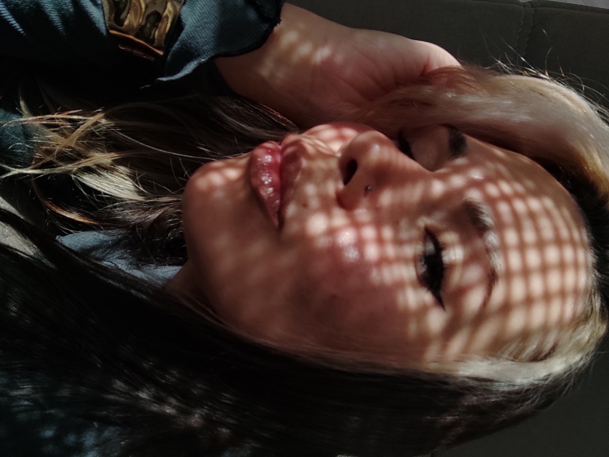 An close-up, high-angle shot captures a person, likely female, reclining indoors, with direct sunlight casting a distinctive grid pattern of shadows across her face. Her eyes are closed or heavily lidded, and her lips are slightly parted, revealing some texture. A small nose piercing is visible on her right nostril. Her hair, a mix of dark and lighter blonde or brown streaks, frames her face and flows around her head. One hand is partially visible, seemingly resting on her head or hair. She is wearing a dark garment, possibly a sweater or jacket, and a metallic, gold-toned object, possibly jewelry or part of her clothing, is visible near her shoulder. The background is dark and out of focus, suggesting an indoor setting like a couch or bed. The strong, directional light and shadows indicate a sunny daytime, possibly morning or late afternoon. The overall mood conveyed is one of relaxation and tranquility. No text is visible in the image. This scene takes place in Chaikovsky, Russia.