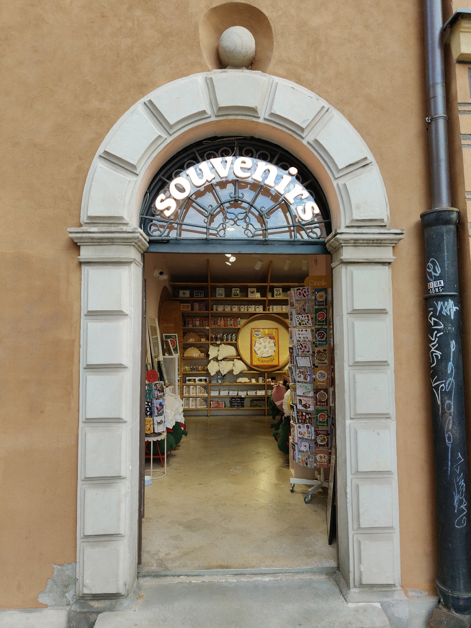 The image shows the entrance to a souvenir shop in Warsaw, Poland. The shop is located under an arched entryway with decorative stone molding. A sign above the entrance reads "Souvenirs" in illuminated lettering. Inside the shop, shelves are stocked with various items, including postcards, small decorative items, and other souvenirs. A man in a green shirt and a man in a pink shirt can be seen in the background. To the right of the entrance is a dark metal pipe with graffiti. The exterior wall of the building is a textured, light brown color.
