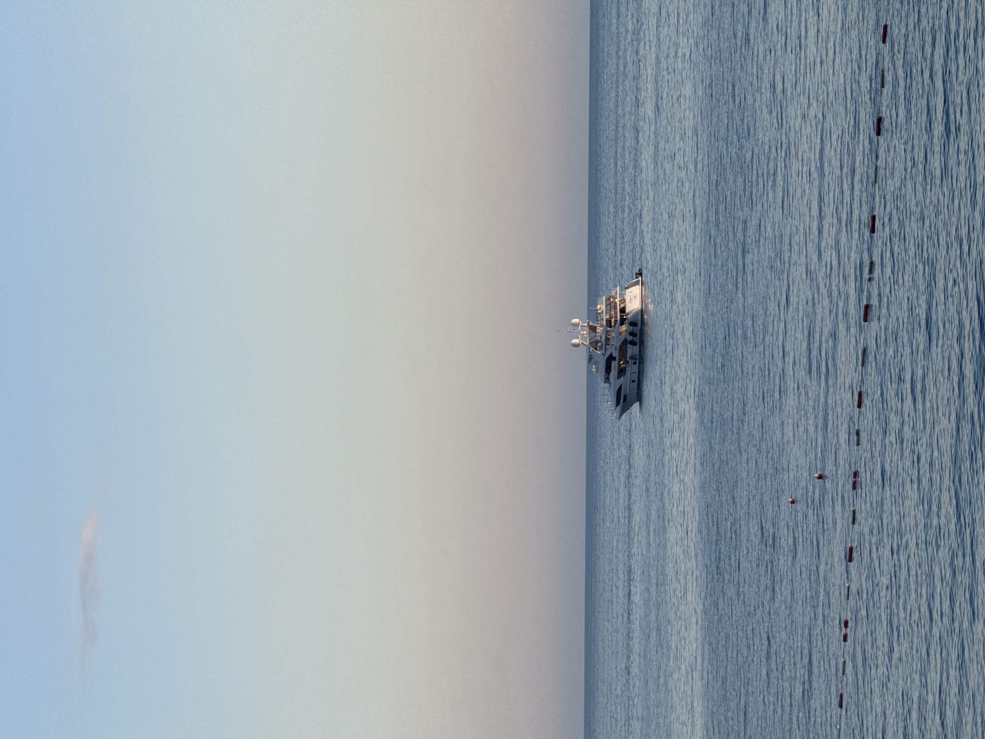 The image shows a white ship on calm blue water under a clear, bright sky, likely near Kamenovo, Montenegro, though the location cannot be definitively confirmed by the image alone. A string of orange and green buoys extends from the right side of the ship. A faint cloud is visible in the sky to the left of the ship.