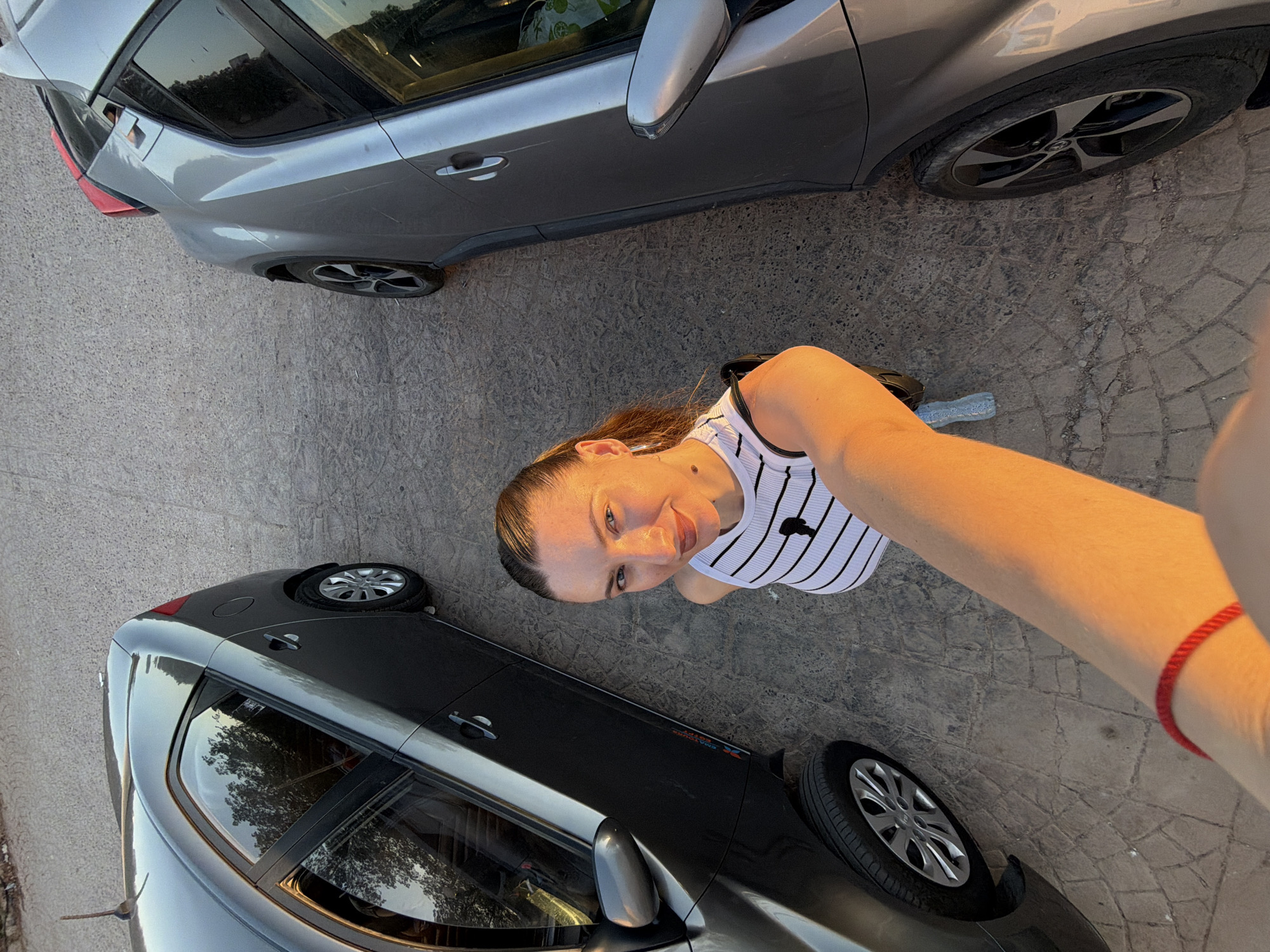 A woman stands between two parked cars in an outdoor setting, likely a parking lot, with a cobblestone surface. She is taking a selfie, with her arm extended towards the camera. The woman has dark hair pulled back and is wearing a black and white striped tank top. She has on a red bracelet on her wrist. The cars are dark grey and silver, and appear to be modern sedans. The lighting suggests it is late afternoon or early evening, with the sun casting a warm glow on the scene. The overall environment is flat and appears to be an urban or suburban area.