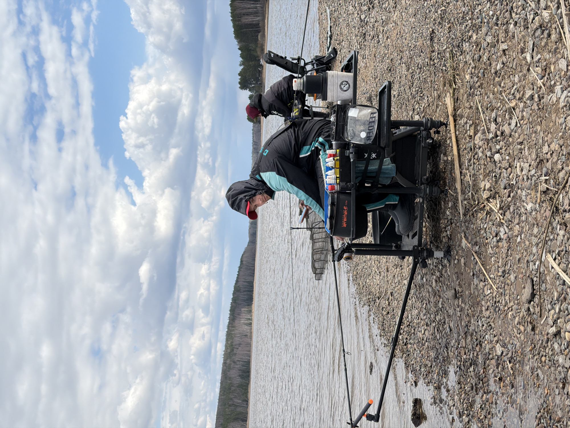 This outdoor scene captures two individuals engaged in feeder fishing on a rocky shoreline by a large body of water, likely in Chaikovsky, Russia.
In the foreground, slightly to the left, a person wearing a dark jacket with light blue panels and a red cap is positioned next to a specialized fishing seat or station. This station is equipped with various fishing accessories, including bait buckets, tackle boxes, and rod holders. Text visible on the station includes "LEEDER," and possibly "WANNER" on the jacket of the person. One bucket features a logo with a fish silhouette, and "3.0 t" is seen on another piece of equipment. A fishing rod extends from the station towards the water.
Further back and slightly to the left, a second individual, also in dark fishing attire, is visible with their own fishing setup, facing the water.
The right side of the image features a calm, wide expanse of water, possibly a lake or a broad river, which reflects the partially cloudy sky. On the distant bank, a dense treeline of evergreen forest stretches horizontally, indicating a natural, unpopulated environment. The immediate shoreline is composed of small, greyish-brown pebbles and gravel, interspersed with dry grasses and reeds.
The sky above is partly cloudy, with patches of clear blue visible through the white, fluffy clouds, suggesting fair weather during the daytime. The overall impression is one of focused fishing activity in a serene natural setting.