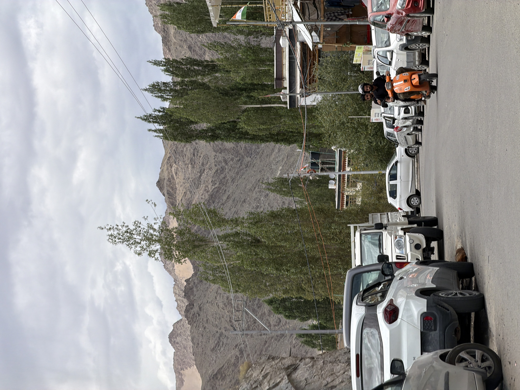 The image captures a street scene in Leh, India, with a backdrop of rugged, dry mountains under a cloudy sky. In the foreground, several cars are parked along the side of the road, including a white SUV, a pickup truck, and a silver sedan. Further down the street, an orange scooter with two riders, both wearing helmets, is in motion. The rider in front is wearing a black shirt, and the passenger behind him is wearing a dark t-shirt. To the right of the scooter, a red car is visible, and behind it, a building with an Indian flag fluttering from a balcony. Tall, slender trees line the street, interspersed with utility poles and wires. The overall atmosphere suggests a typical day in a mountainous town.