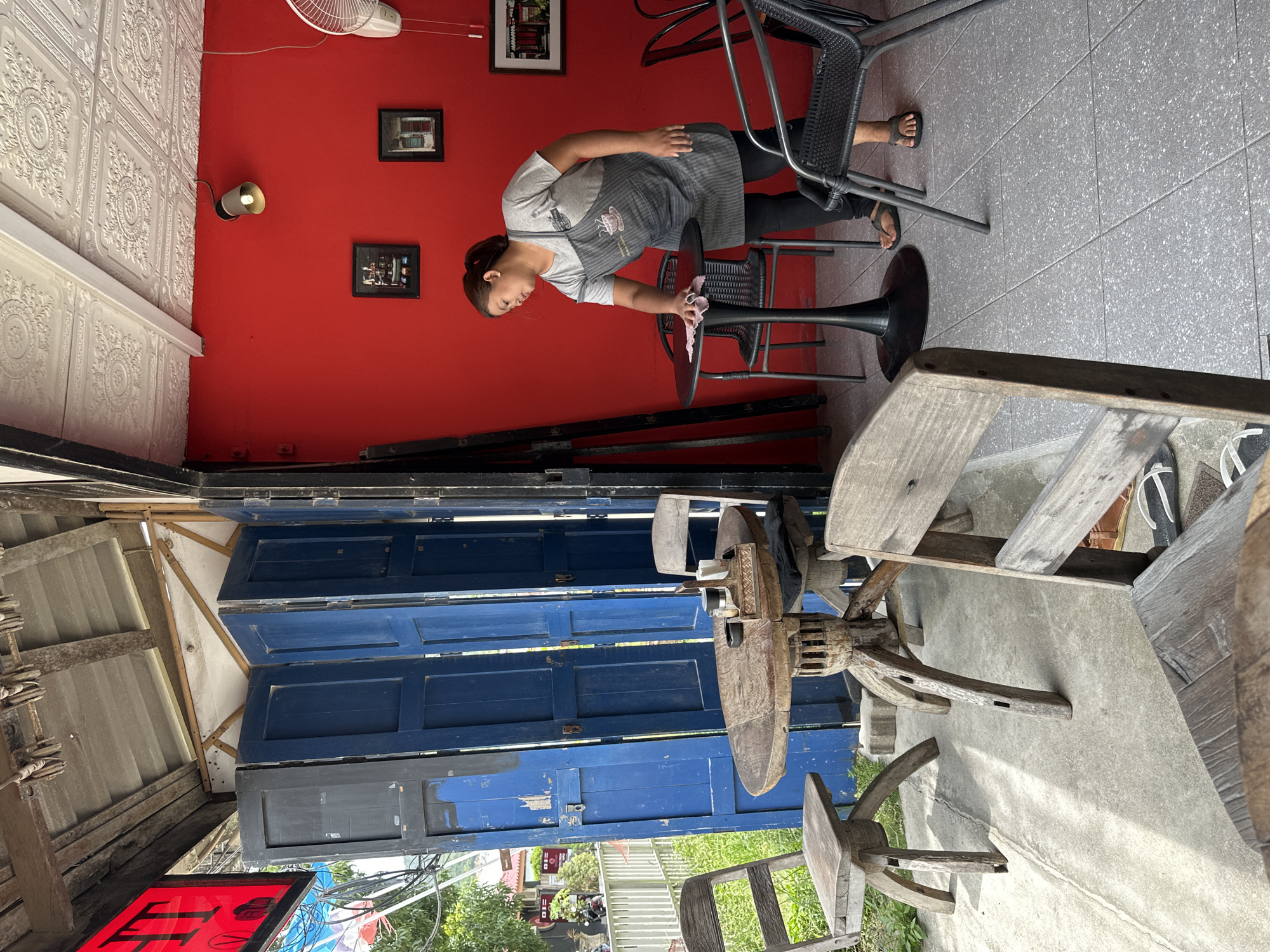 The image captures a cafe or restaurant setting, featuring both an indoor and an outdoor seating area. The main subject is a woman, likely a staff member, wearing a grey t-shirt, dark pants, and a grey striped apron with a coffee cup design. She is actively wiping a small, round black table with a pink cloth, standing next to a black metal bar stool. She wears open-toed sandals.
The indoor area is characterized by a vibrant red wall adorned with two framed pictures and a speaker/light fixture. A white ceiling fan is visible near the top left corner of the red wall. Adjacent to the red wall is a distinct white wall or ceiling panel with an intricate, embossed pattern. The floor in this section is composed of grey tiles.
Transitioning from this area are several large, distressed blue wooden doors, which appear to be used as a partition or decorative element, adding a rustic touch. Beyond these doors lies what seems to be an outdoor section, featuring a concrete ground and a collection of rustic wooden tables and chairs/stools, some with distinct carved bases. Green foliage and parts of an outdoor structure or fence are visible in the background, suggesting a natural outdoor environment.
The lighting is bright, indicating it is daytime. A red sign with partially visible white text, possibly stylized characters or Thai script, is seen in the very bottom left corner but is not clearly legible. The scene combines modern elements, like the bright red wall, with a more traditional or rustic aesthetic from the wooden doors and outdoor furniture.