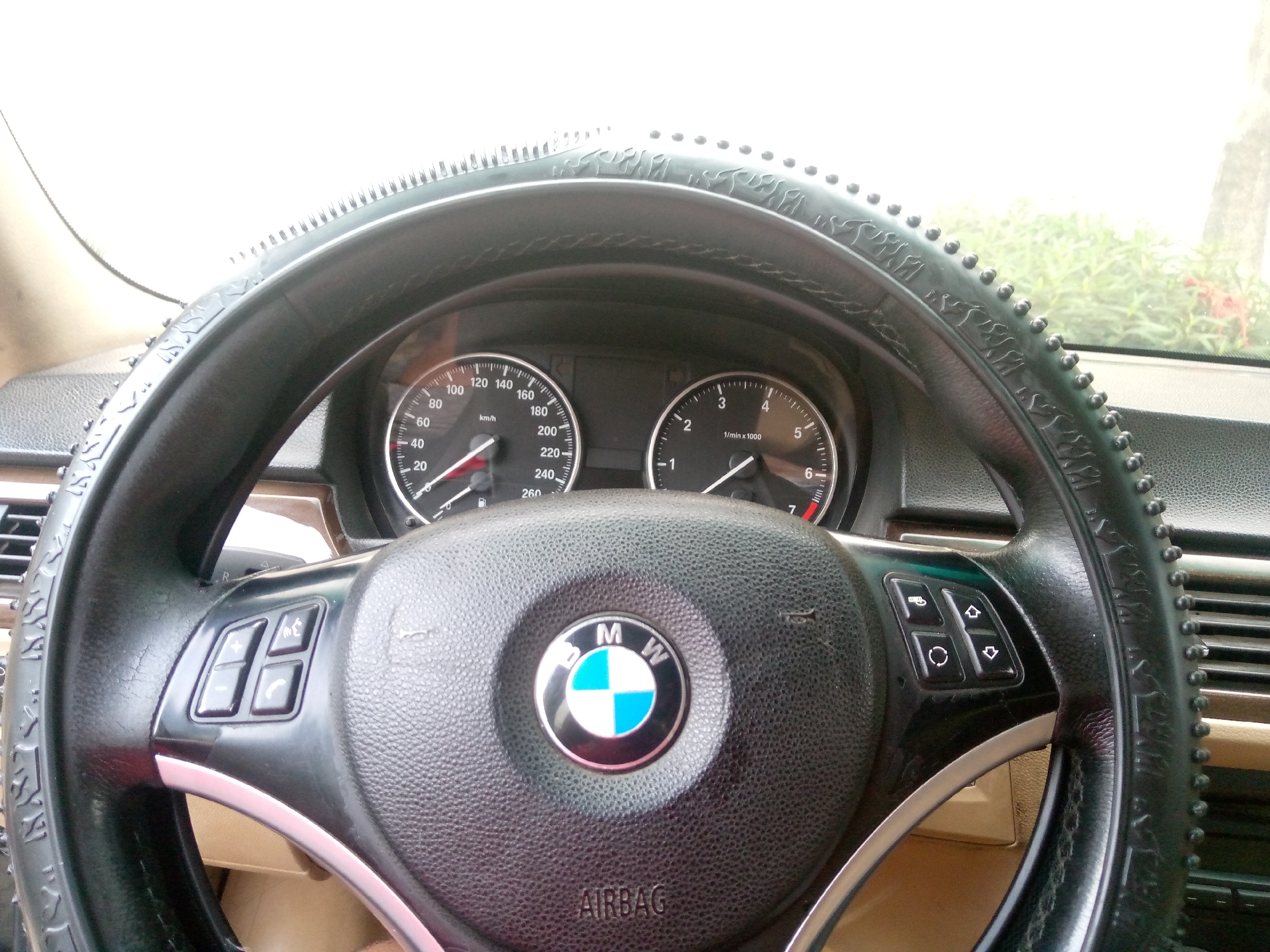 The image shows the interior of a BMW car, focusing on the steering wheel and dashboard. The steering wheel is black and has a textured cover with raised dots along the outer rim. The BMW logo is prominently displayed in the center of the steering wheel. To the left and right of the steering wheel are various buttons for controlling the car's functions, including volume, calls, and possibly other infotainment features. The dashboard includes two main gauges: a speedometer on the left, showing speeds up to 240 km/h, and a tachometer on the right, measuring engine revolutions per minute. The car's interior appears to be in moderate condition, with some visible wear on the steering wheel cover. The background suggests the car is parked, with greenery visible through the windshield, hinting at an outdoor location. The image context provided indicates the location is Jalingo, Nigeria.