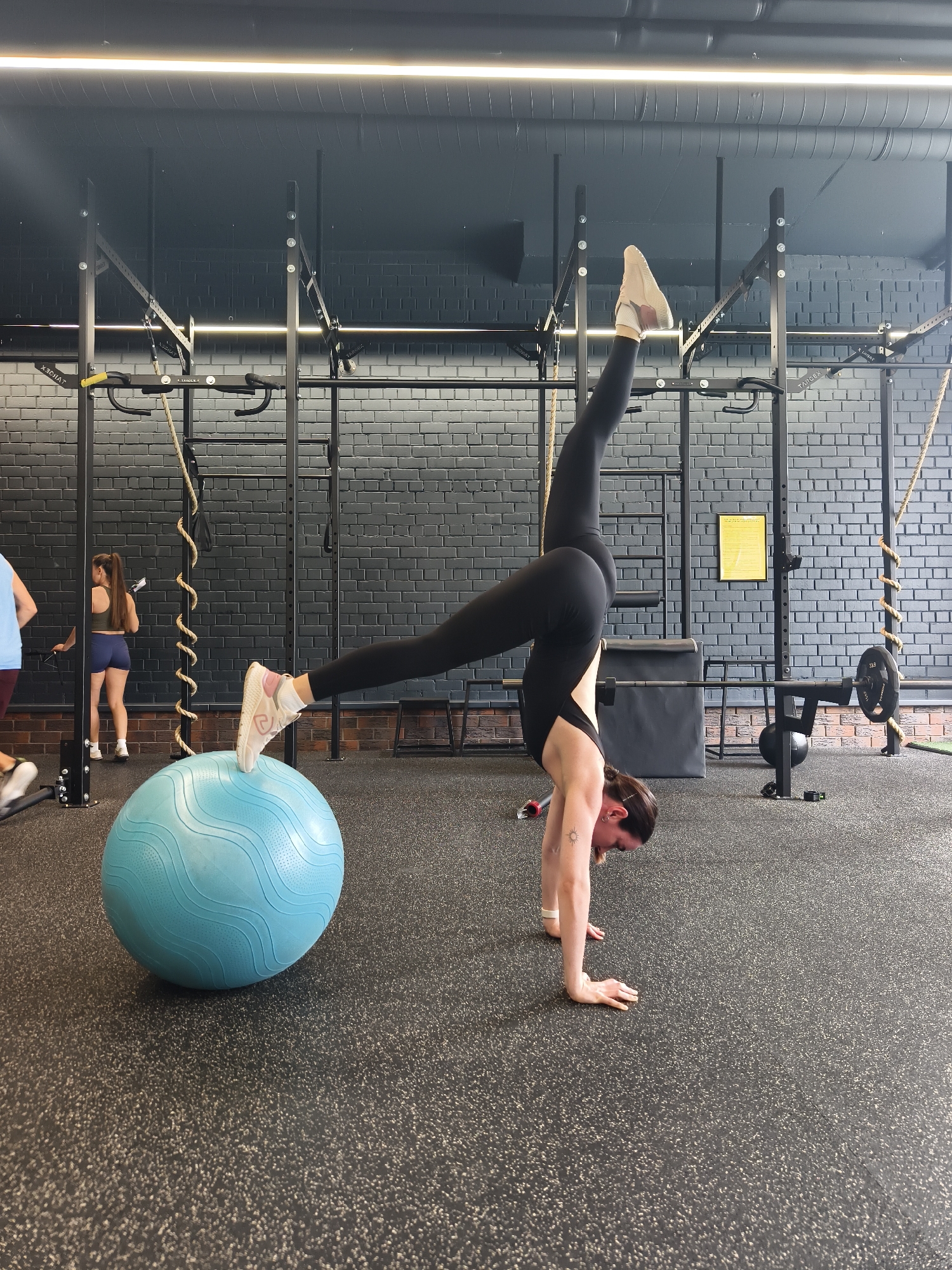 This image depicts an indoor gym or functional fitness facility in Tyumen, Russia. The central subject is a woman performing a handstand exercise. She wears black athletic leggings and a matching sports top, with light-colored athletic shoes. Her hands are on the speckled black rubber gym floor, while her left foot is balanced on a large, light blue stability ball, and her right leg is extended vertically towards the ceiling. A small sun tattoo is visible on her upper right arm, and she appears focused on maintaining her balance.
In the background, two other individuals are visible. On the far left, a person wearing a blue shirt and dark shorts is partially seen. Further back, near a black metal rig, another woman in an olive-green top and blue shorts stands with her back to the camera.
The gym features black brick walls and a dark ceiling with exposed pipes and bright linear fluorescent lights. Essential gym equipment includes a robust black metal rig with pull-up bars and a thick climbing rope. Text such as "X30HAT" (reversed, likely a brand name) and "MAROOX" can be seen on the rig. To the right, a barbell with a "30 LB" weight plate is visible. A yellow rectangular sign hangs on the far wall. The overall ambiance suggests an active training session in a well-equipped, modern fitness environment.