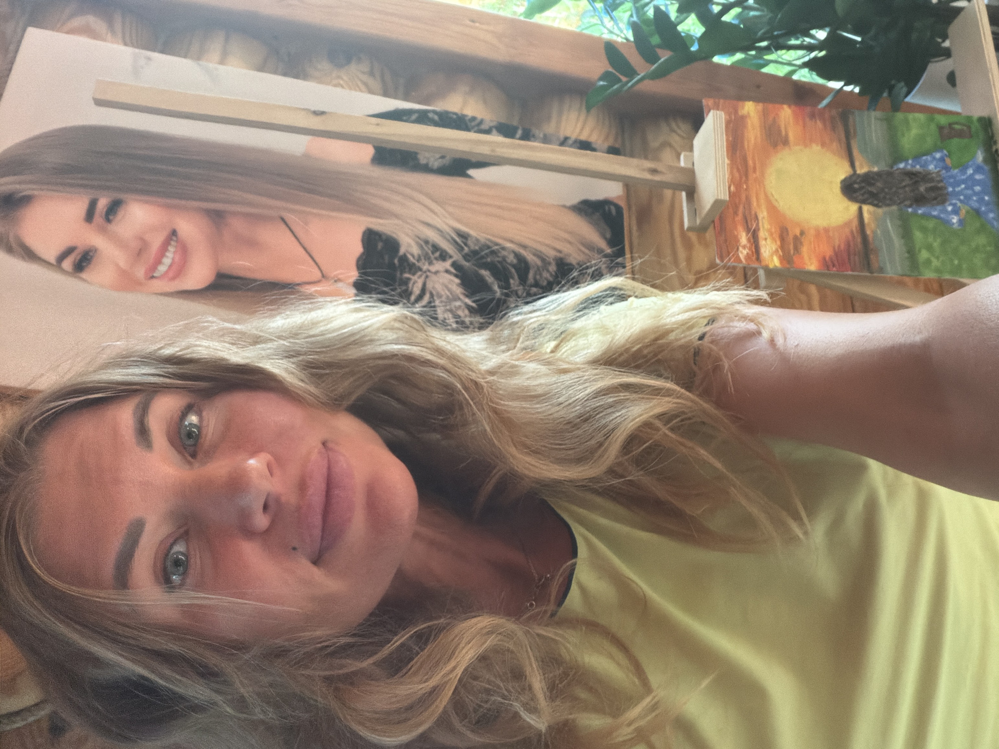 A woman with blonde, wavy hair and blue eyes is taking a selfie. She is wearing a yellow t-shirt and a necklace. In the background, there is a large painting of another woman with long blonde hair and a black dress, and a smaller painting of a woman with long brown hair wearing a blue dress, looking at a sunset. The scene appears to be indoors, possibly an art studio or a home. There are plants and wooden structures visible, suggesting a natural or rustic environment. The image does not provide any information that would allow for the identification of the specific location as Tagayevo, Russia.