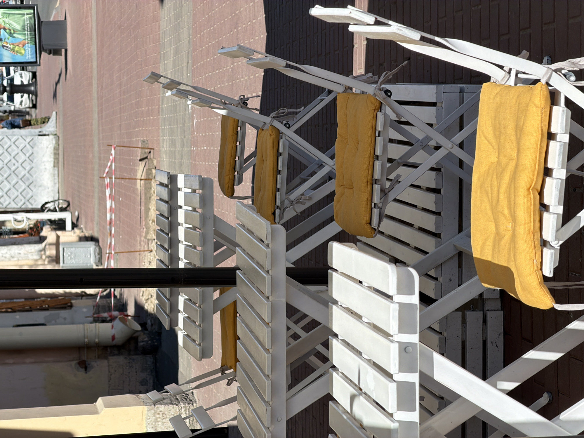 This full-frame, eye-level shot depicts an empty outdoor cafe seating area on a sunny day in Saint Petersburg, Russia. In the foreground and midground, four sets of white, slatted, foldable wooden tables and chairs are arranged on a reddish-brown brick-patterned pavement. Each chair is fitted with a bright yellow fabric cushion, and strong sunlight casts defined shadows from the furniture across the ground. A dark, vertical pole, likely part of an umbrella stand, is positioned centrally among the tables.
In the background to the left, a segment of a light-colored building facade is visible, featuring a prominent white downspout that descends to ground level. A small area near the building is cordoned off with red-and-white striped warning tape. To the right, the pavement extends into a wider street or pedestrian area where several cars, including a white sedan, are visible in the distance. A dark lamppost stands near the street, and a large, illuminated billboard displays an advertisement featuring a person holding a green bottle against a blue background, accompanied by text in Cyrillic script. Distant figures of people are discernible further down the street. The Cyrillic script on the billboard and the architectural style of the buildings are consistent with an urban environment in Saint Petersburg, Russia.