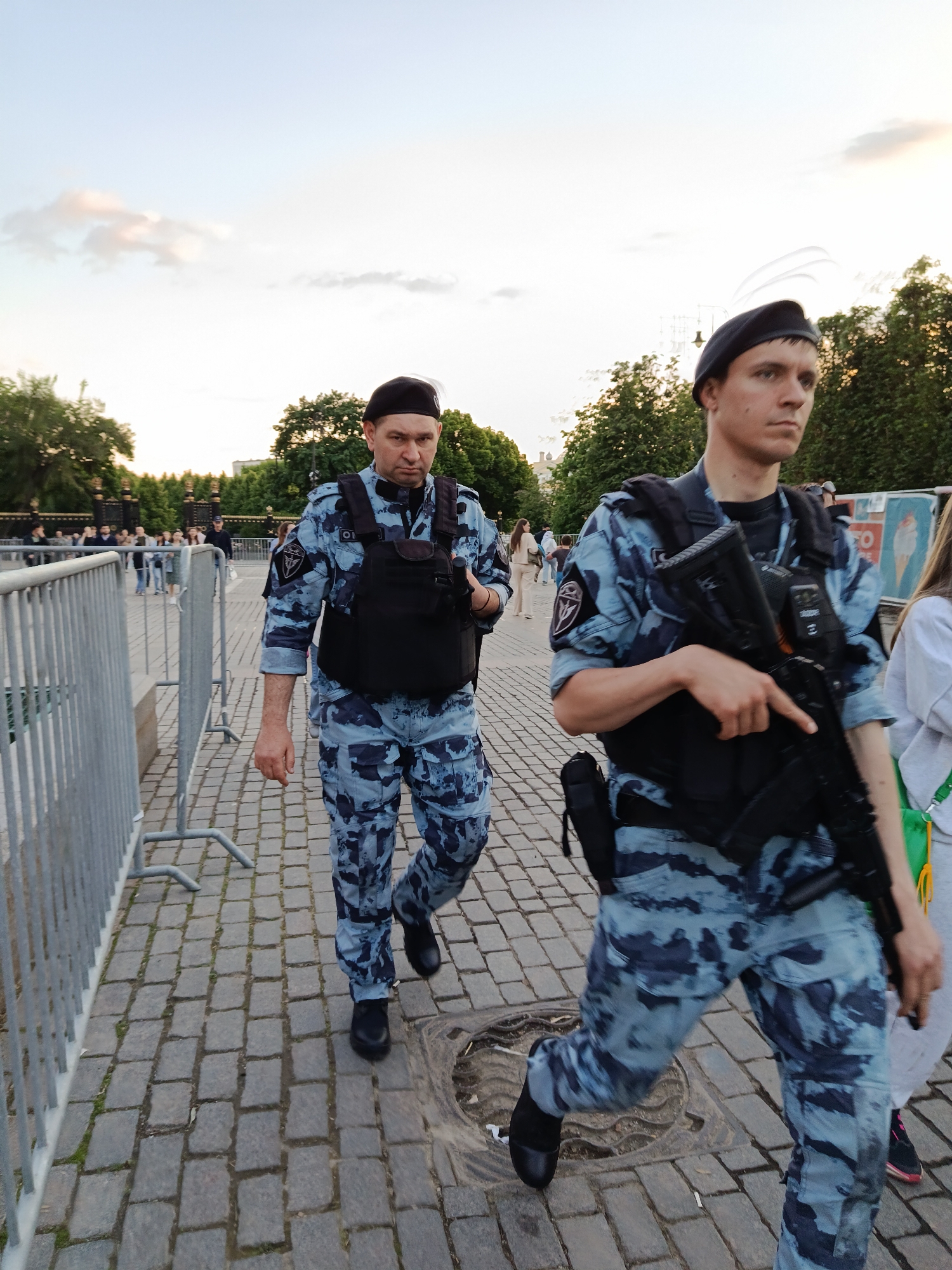 Two men in light blue and grey digital camouflage uniforms, black tactical vests, and black berets are walking outdoors on a paved area. The man in the foreground on the right carries an assault rifle slung across his chest. Both men have shoulder patches featuring a circular emblem, consistent with Russian security forces like Rosgvardia. The man on the left has what appears to be "O.L." or similar initials on a small patch on his vest.
They are walking alongside a grey metal crowd control barrier on a cobblestone pavement. In the background, other people can be seen, and beyond them are green trees and some ornate gates or fences, suggesting a public park or square. The time of day is late afternoon or early evening, indicated by the soft, ambient light and a bright sky with faint clouds. The men's expressions are serious, suggesting they are on duty or patrol. On a partial sign in the distant background to the right, the word "ICE" is visible. The ground is a distinct cobblestone pattern, and a circular manhole cover is visible near the feet of the man on the right.