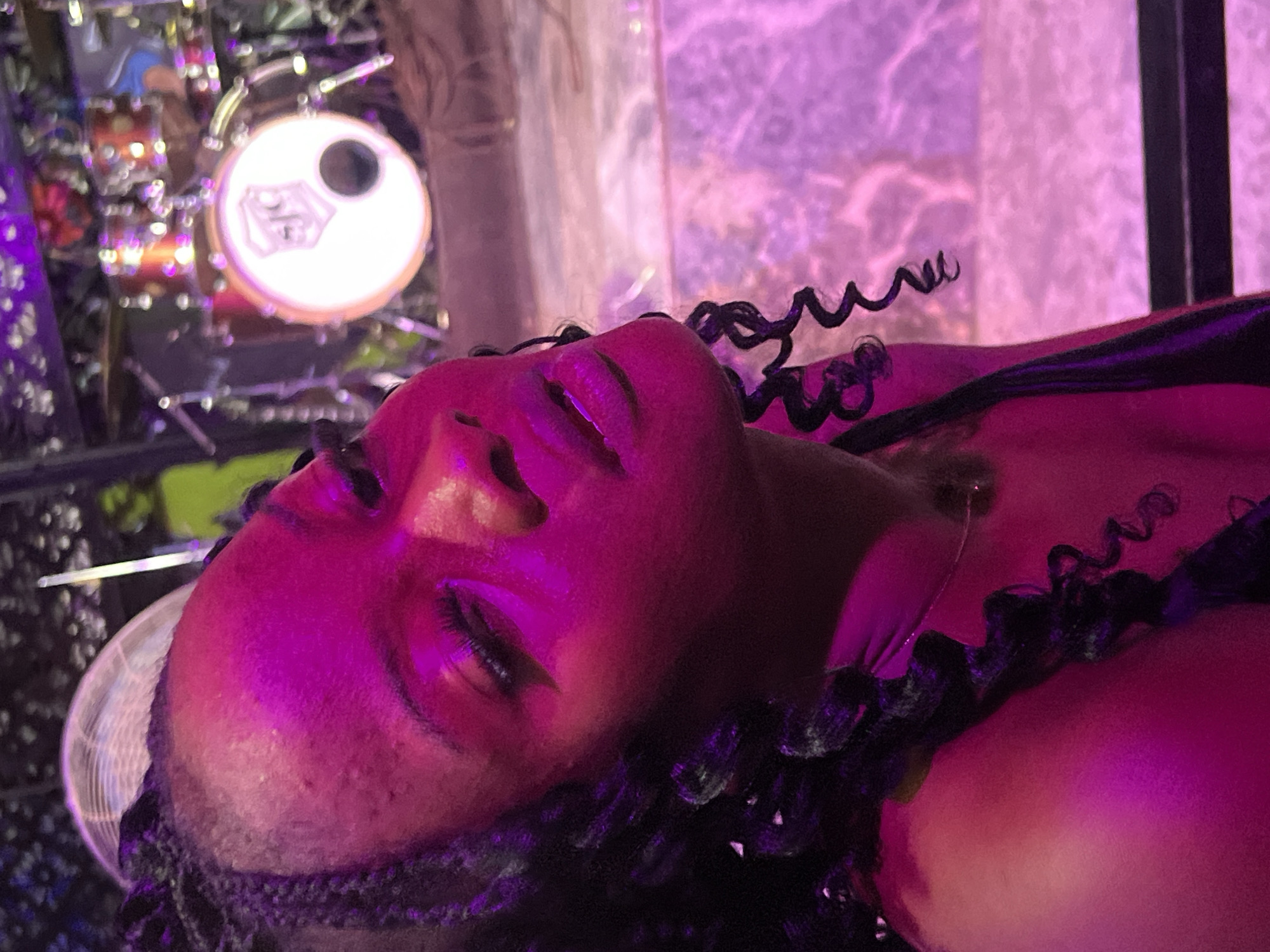 A close-up, low-angle shot reveals a woman lying down. The scene is illuminated with a magenta light. A drum set with a prominent white drumhead sits blurred in the background, while a fan is visible on the left side of the frame. The woman is adorned with curly black hair. She is possibly located in Howayi, Nigeria.