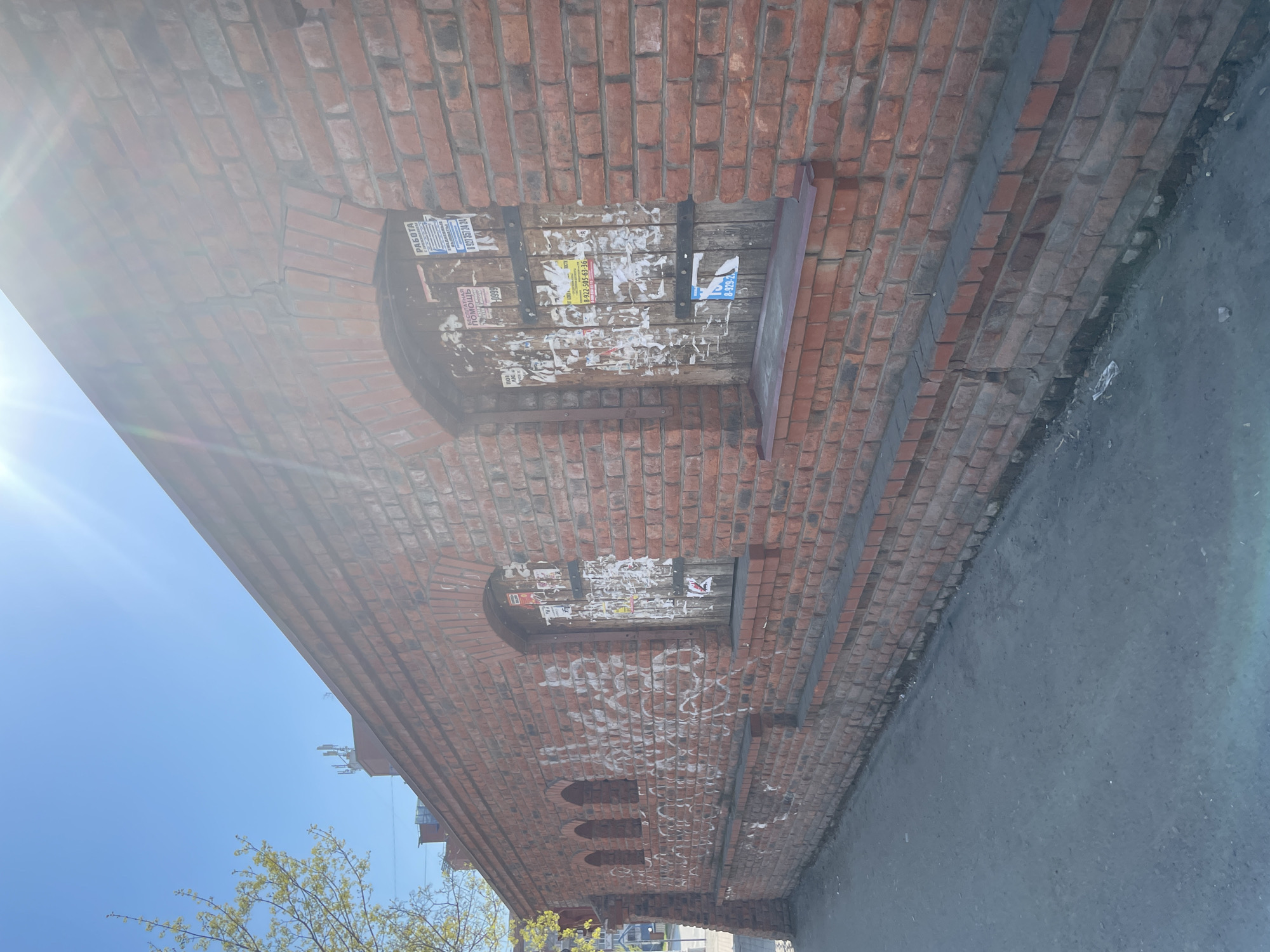 The image shows a brick building in Izhevsk, Russia. The building has arched windows, some of which are boarded up and covered with posters. There is graffiti on the brick wall. The building is located next to a street. The sky is visible in the background. A tree with green leaves is in the lower left corner.