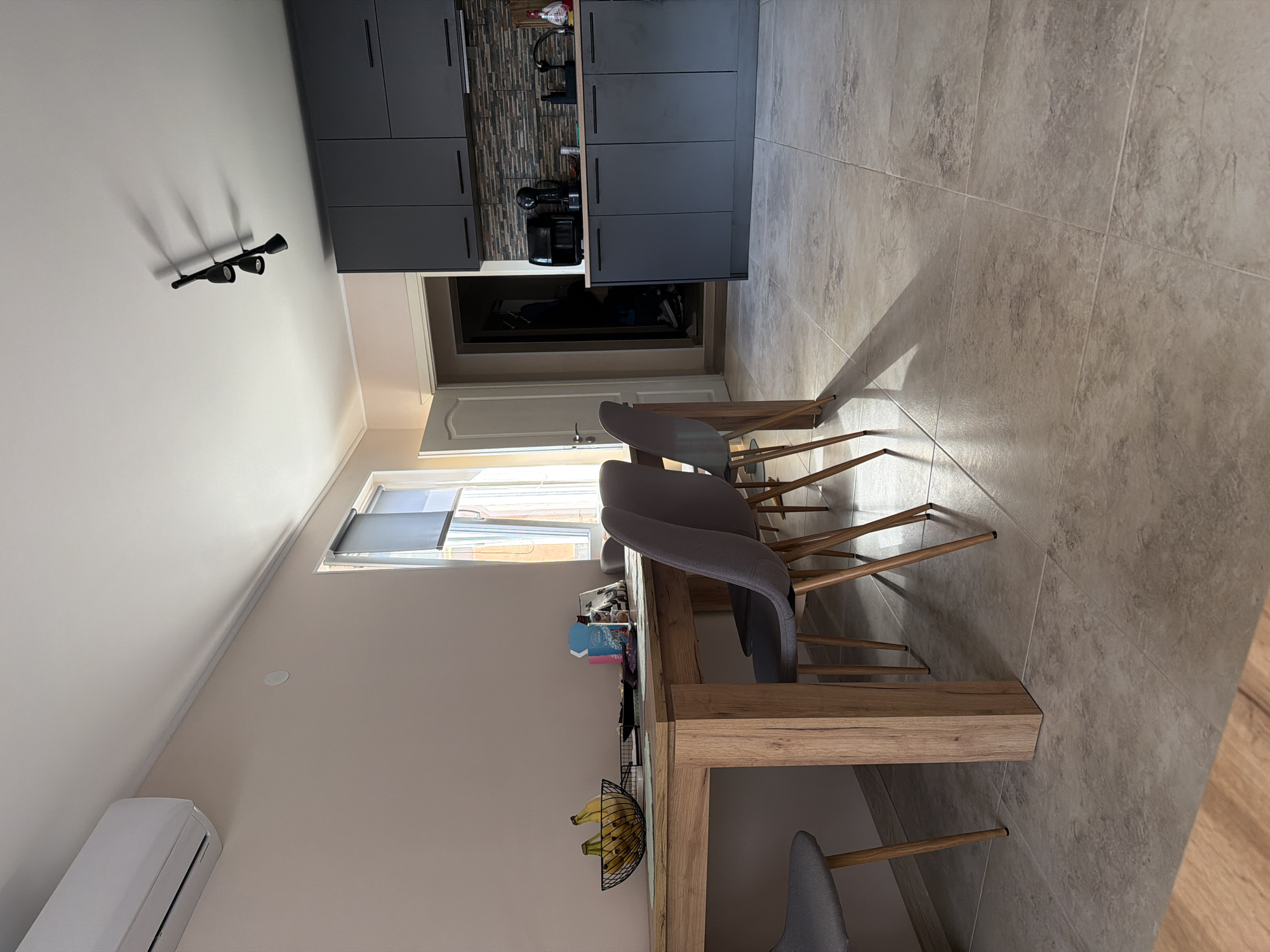 This is an interior shot of a kitchen and dining area. The kitchen features dark gray cabinets and a tile backsplash. There is a doorway visible, and a wall-mounted air conditioning unit is present. The dining area includes a light wood table with gray chairs. A fruit basket with bananas is on the table. A roller blind is visible on the window. The flooring is a light brown tile. The location is Litakovo, Bulgaria.