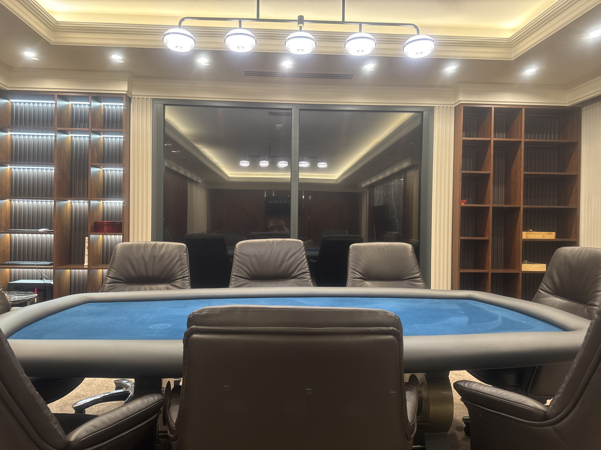 A poker table is the focal point of the image, surrounded by leather chairs, likely in a room or parlor with a large reflecting window. The room is lined with built-in shelving. The location is unknown and cannot be determined from the image.