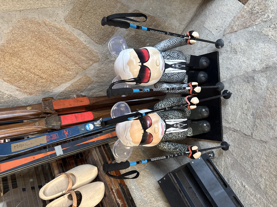 The image depicts an indoor display located in Courchevel, France. Two large, stylized figures resembling Karl Lagerfeld, rendered in a Bearbrick-style with translucent ears, stand prominently on black bases. Both figures wear black sunglasses, feature white hair, and are dressed in grey patterned jackets over black shirts with white neck detailing. Each figure grasps two black and blue Rossignol ski poles with black handles and orange/pinkish straps, positioned with their baskets touching the flagstone floor. In the background, propped against a rustic stone wall composed of irregular tan-brown blocks, are several pairs of skis: a set of older wooden skis with red tips and a pair of blue and orange Rossignol skis, one bearing a "PLAGNE" sticker. To the left of the skis, two pairs of light wooden clogs or traditional shoes rest on a dark wooden surface. A black metal stand is partially visible on the far left. The floor consists of large, irregular grey flagstones.