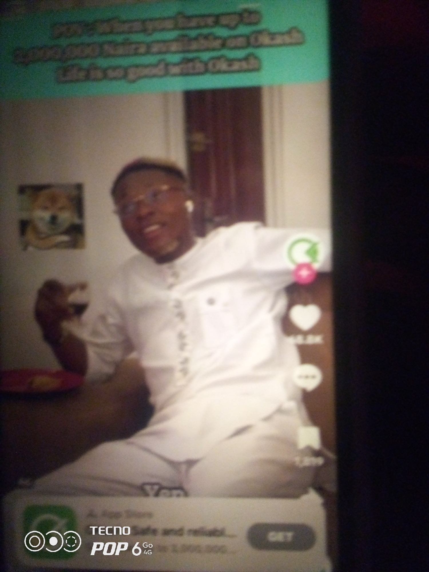 The image shows a close-up of a person, likely male, wearing a white garment with intricate silver embroidery on the front. He appears to be in an indoor setting, possibly a living room. He is holding a small glass in his right hand. To his left, a framed picture of a dog is visible on the wall. The background is somewhat blurred, but it appears to be a wall. Overlaying the scene are graphical elements commonly found on social media platforms, including a green circular icon with a refresh symbol, a pink circular icon, a heart icon with a number "5.8K" below it, and a bookmark icon with the number "1,319". At the bottom of the image, there is a branding element for "TECNO POP 6 4G" with a button labeled "GET". The text at the top of the image, partially visible, reads "POV: When you have up to 1,000,000 Naira available on Okash. Life is so good with Okash". There is no visible indication of the specific location within Howayi, Nigeria, in the image itself.