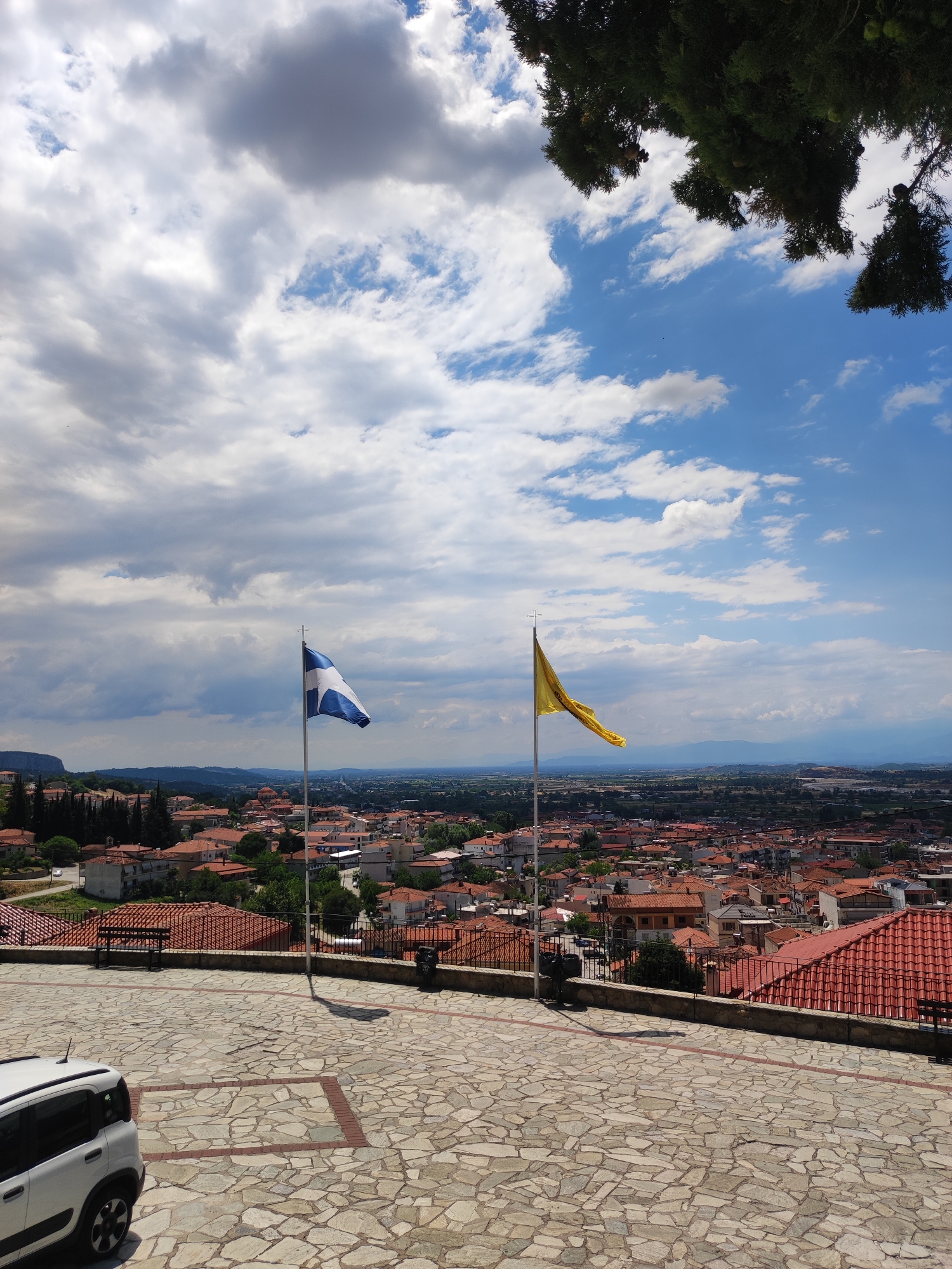 The image depicts a scenic overlook in Kalabaka, Greece, featuring two flags on flagpoles. The flag on the left is blue and white with a cross, likely the Greek flag. The flag on the right is yellow and appears to have an emblem, possibly a religious or regional flag. Both flags are elevated, with the Greek flag slightly to the left of the yellow flag, and they are positioned against a backdrop of the city's rooftops and a distant landscape.
The foreground is dominated by a paved courtyard made of irregular stone tiles. To the left, a white car is partially visible, parked facing towards the right. A dark-colored bench is situated along a low stone wall that borders the courtyard, overlooking the city. Beyond the wall, the dense urban landscape of Kalabaka unfolds, characterized by numerous buildings with red-tiled roofs. Lush green trees are interspersed throughout the city and on the surrounding hills.
The sky is a mix of blue patches and dramatic white and grey clouds, suggesting dynamic weather conditions. The overall impression is one of a bright, possibly windy day with a commanding view of the town and its surroundings. The time of day appears to be midday or early afternoon, based on the lighting and shadows. The presence of the flags suggests a location of significance or a public space.
