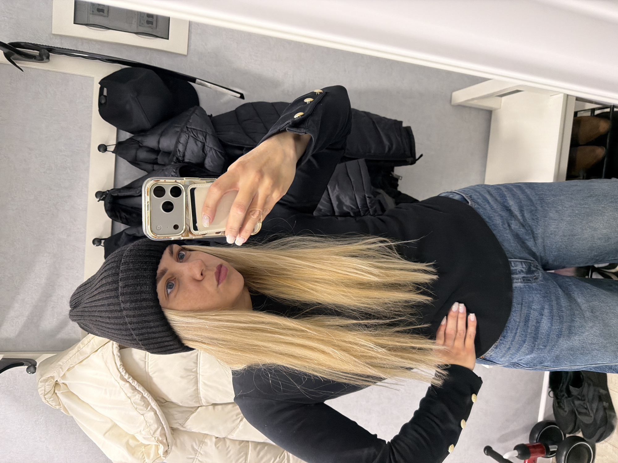 A woman is taking a selfie in what appears to be a home or entryway. She is wearing a black beanie, a long-sleeved black top with gold buttons on the sleeves, and blue jeans. Her long, blonde hair is down. She's holding a phone in her right hand, taking the picture. Behind her, there is a rack with several coats hanging on it: a black down jacket, a black baseball cap, and a cream-colored puffer jacket. A white shelf unit is also visible, as well as a glimpse of what might be shoes. A painting or picture is hanging on the wall above the rack. There is a child's tricycle on the floor. The setting doesn't suggest any specific location within Votkinsk, Russia.