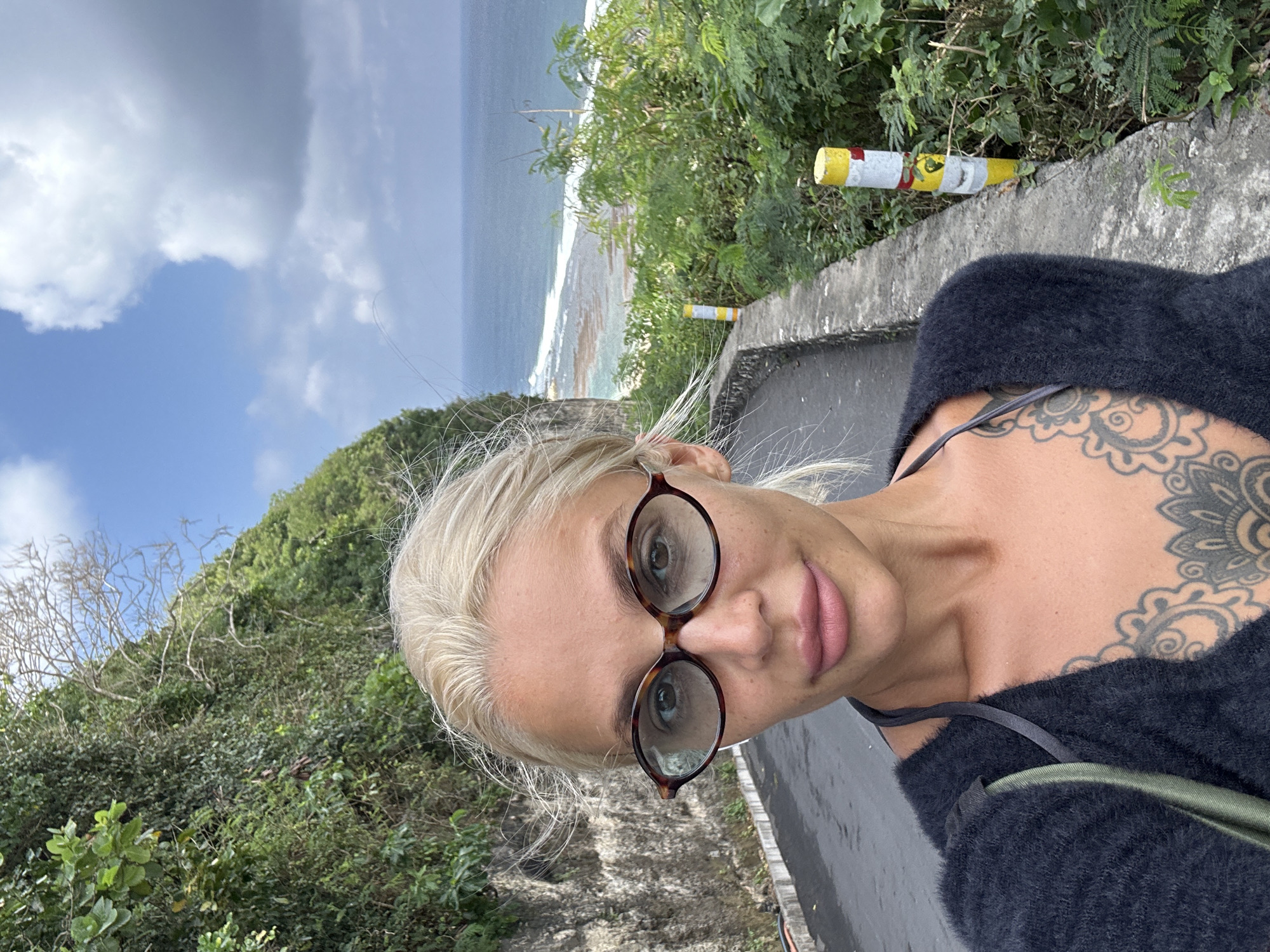 A woman with blonde hair and tattoos on her chest takes a selfie on a paved road with lush green foliage and the ocean in the background. She is wearing a dark, fuzzy sweater and oval-shaped glasses with tortoise frames. The road appears to be winding uphill. Two yellow and white striped posts are visible along the side of the road. The ocean is visible in the distance with waves breaking on a rocky shore. The sky is blue with scattered clouds. The overall impression is a scenic overlook in a tropical location.