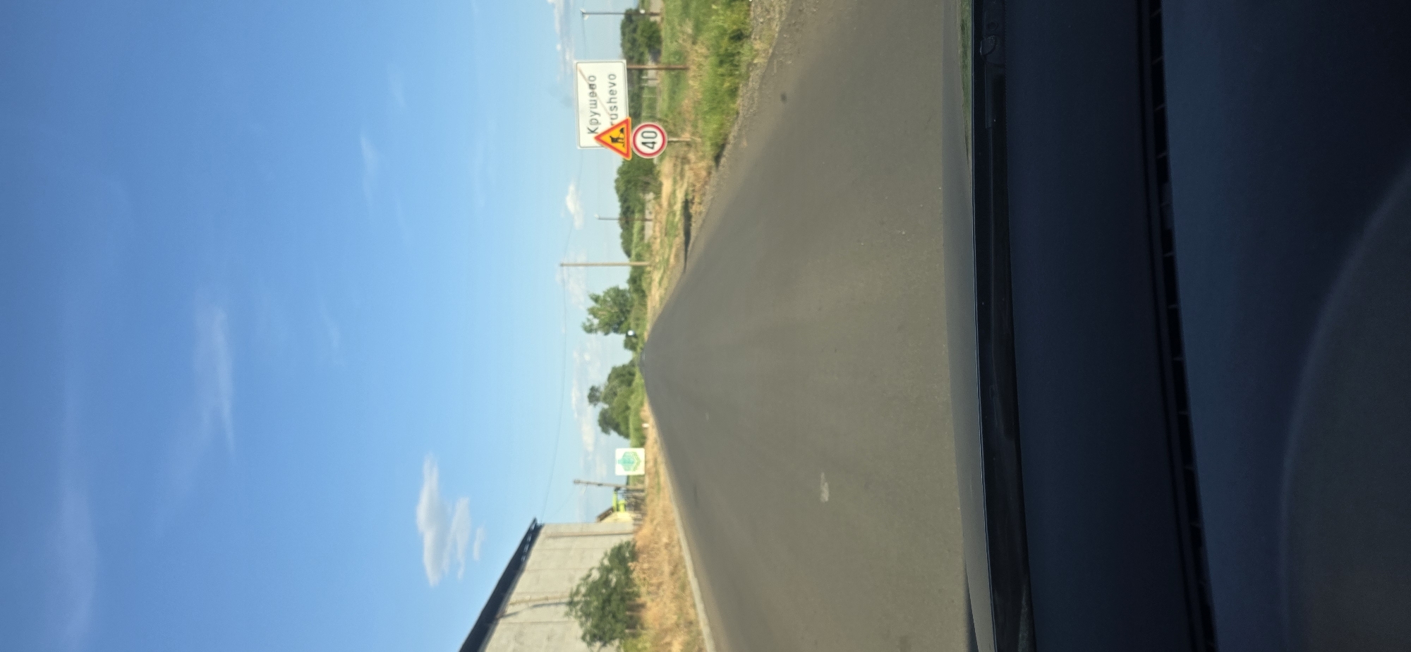 The image shows a road in the city of Krushovo, Bulgaria. A sign indicates the name of the city in Cyrillic ("Крушево") and Latin script ("Krushevo"). A triangular warning sign depicting children is visible, accompanied by a circular speed limit sign displaying "40." The road surface appears to be asphalt. The sky is blue with some scattered clouds. The perspective is from inside a vehicle, with a portion of the car's interior visible on the right side of the frame. Vegetation lines the sides of the road. A building can be seen to the left of the road. Power lines are present.