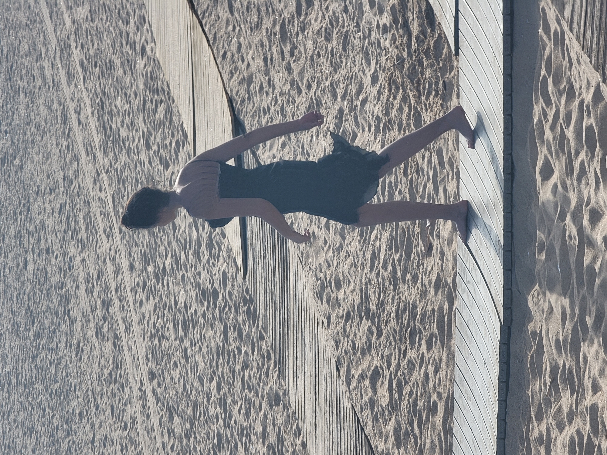 This is a high-angle, full shot of a person lying on a beach boardwalk in Quarteira, Portugal. The person, viewed from above, is dressed in a dark, knee-length dress. Their arms are extended, with the right arm bent at the elbow and the left arm stretched out. Their legs are also extended. The person appears relaxed, with their head resting on the boardwalk.
The setting is a sandy beach, with the texture of the sand clearly visible, showing ripples and footprints. A wooden boardwalk runs diagonally through the frame, with light-colored wooden planks creating a textured surface. The contrast between the rough sand and the smooth wood is evident. The lighting suggests it is daytime, with shadows cast by the boardwalk and the person onto the sand, indicating the sun is at an angle. The overall atmosphere is calm and serene, suggesting a moment of rest or leisure on a sunny day. No text is visible in the image.