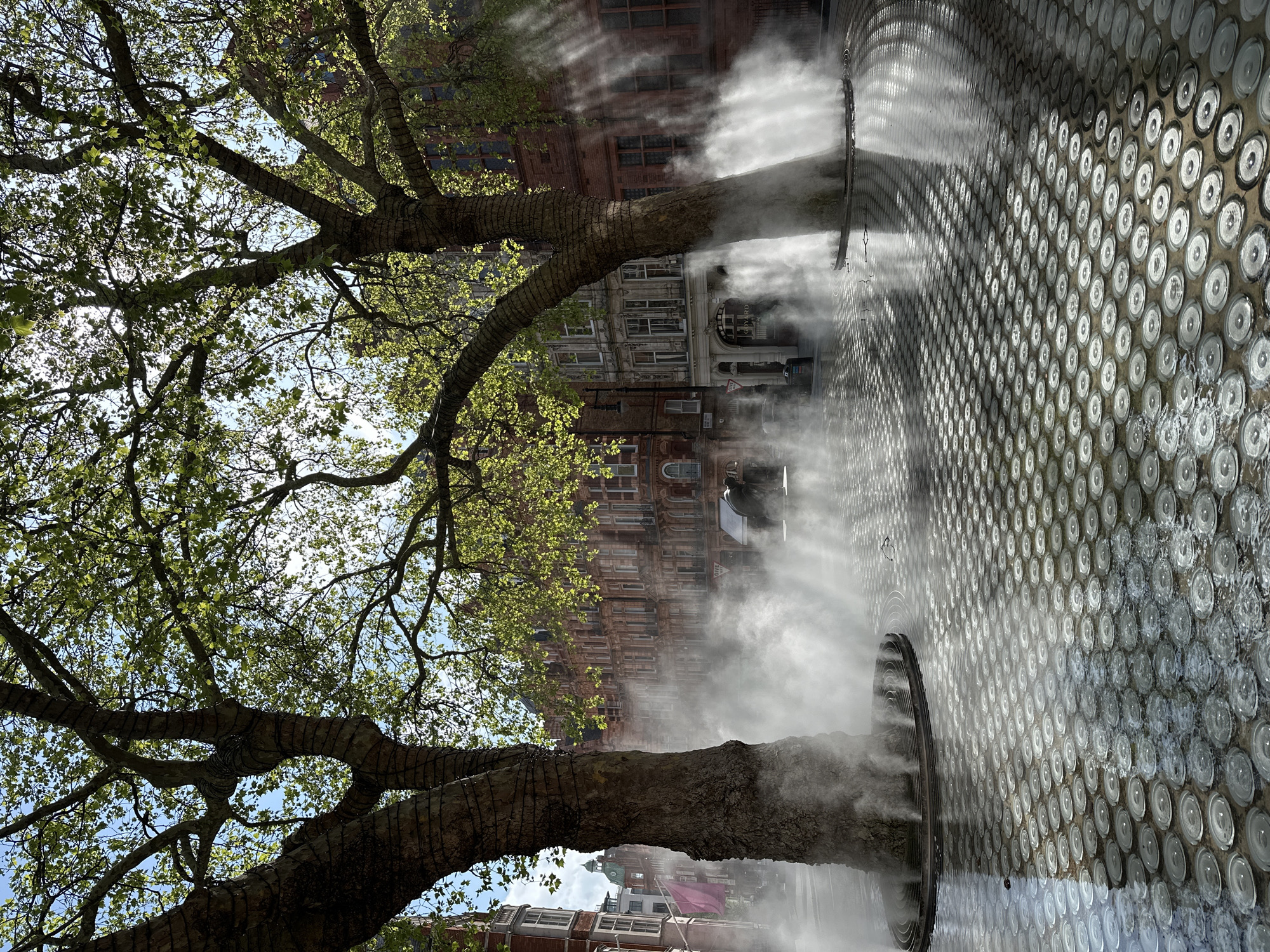 This image captures a vibrant urban scene in London, United Kingdom, under bright daylight. Lush green foliage from two large trees fills the upper and left portions of the frame, with their thick trunks extending downwards. The tree trunks are visibly wrapped, possibly with protective material or decorative lighting.
Rising from the ground on the right side of the image are several plumes of white mist or fog. These emanate from circular, dark installations embedded within a distinctive pavement. This pavement, which dominates the lower right, features a grid of numerous circular, reflective or translucent elements. Some of these circles appear wet or contain water, creating a shimmering, textured surface.
Behind the mist and trees, on the left side of the frame, stand ornate, multi-story brick buildings. Their architectural style, with detailed facades and multiple windows, is characteristic of historic London. Through the leaves and mist, a dark vehicle is faintly visible in the mid-ground. A red "GIVE WAY" road sign is also visible. Part of a building sign shows the letters "FORD".
The clear blue sky is visible through gaps in the tree canopy, suggesting good weather. The overall mood is serene and dynamic, with the bright sunlight illuminating the fresh foliage and the ethereal mist, which likely serves as an artistic installation or a cooling feature.