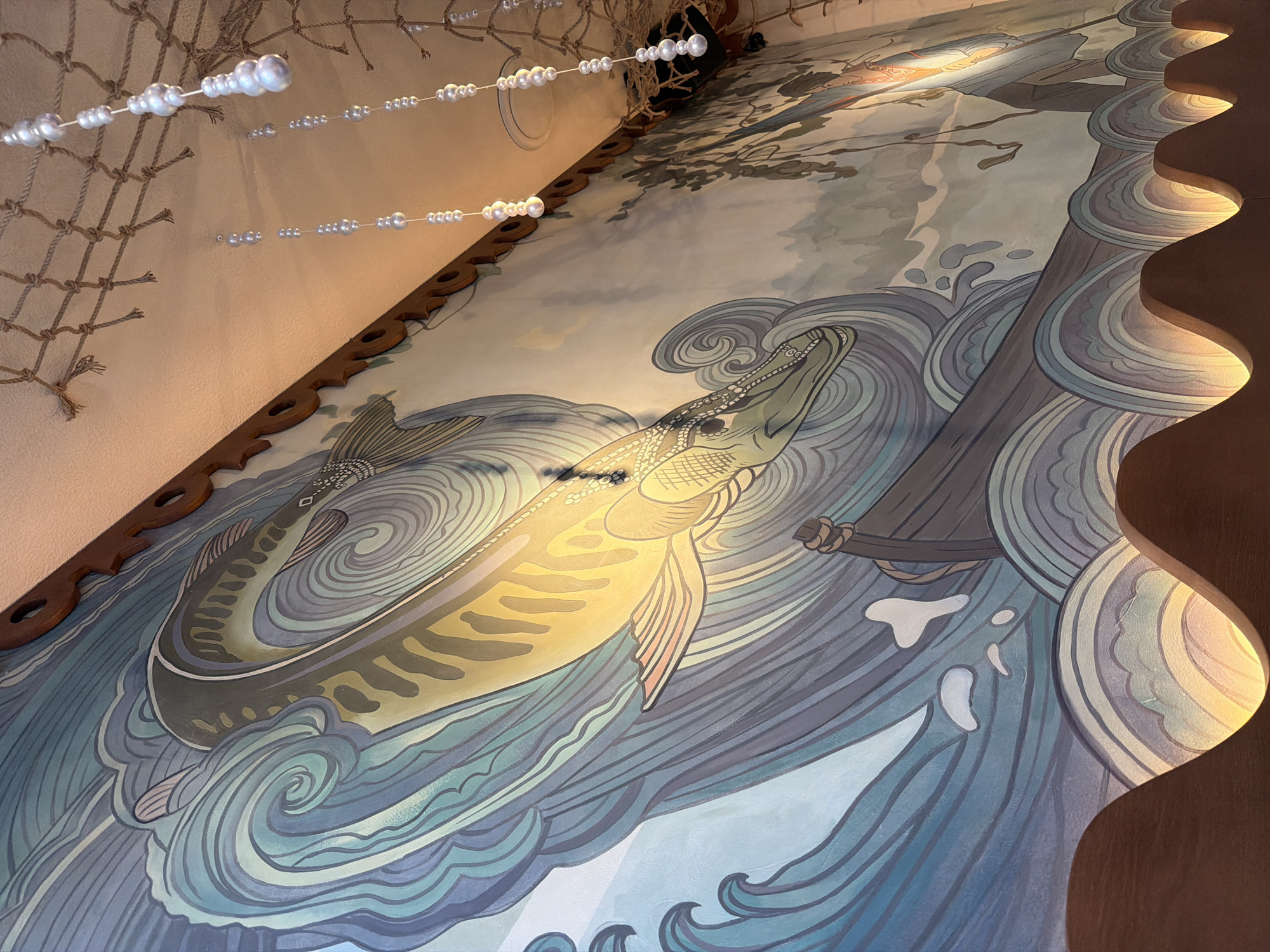 This is a mural painted on a wall, possibly in a restaurant or public space in Nizhny Novgorod, Russia. The mural depicts a stylized scene of a large fish, possibly a pike or musky, swimming through turbulent blue and green waves. The fish is rendered in shades of yellow and green with dark markings. Below the fish, a wooden boat is visible, also amidst the waves. The mural has a folk art or decorative style, with bold outlines and a limited color palette. In the upper right portion of the image, a fishing net is suspended from the ceiling, along with decorative strings of white beads. The ceiling itself is textured and light-colored. The bottom edge of the image shows a wooden structure with integrated lighting that casts a warm glow upwards, creating a wavy pattern on the wall below the mural. There are no discernible people in the mural itself, though a silhouette of a figure can be seen in the background on the right side.