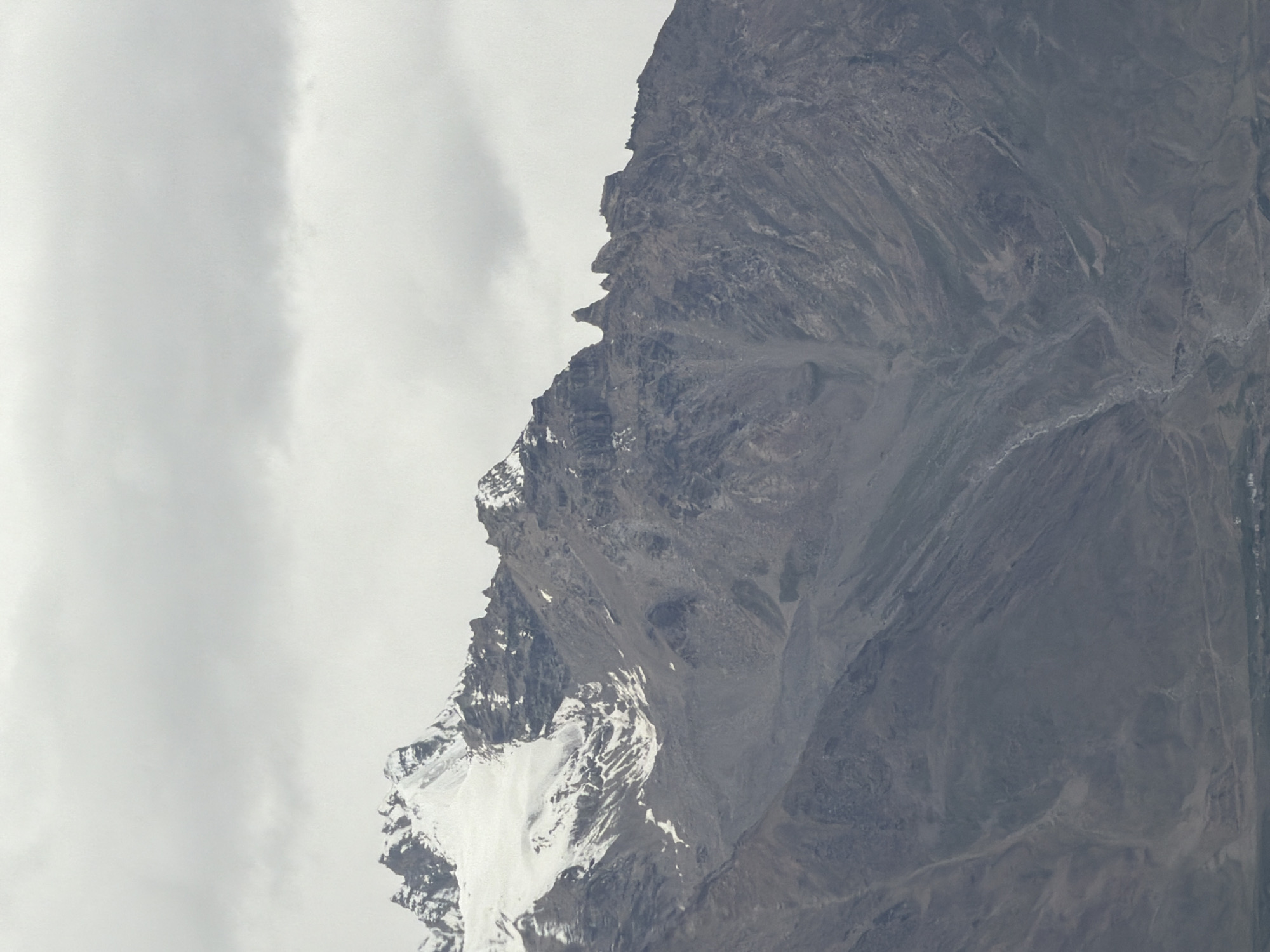 A majestic mountain range under a cloudy sky is captured in this vertical, eye-level shot. The peaks are rugged and dramatic, with snow-capped summits that gleam against the muted gray of the clouds. The mountain slopes are a tapestry of rock and scree, with deep ravines and channels carved into their sides, suggesting a history of erosion and geological activity. In the distance, a small settlement is visible at the base of the mountains, a cluster of buildings suggesting human habitation in this remote landscape. The overall impression is one of vastness, natural power, and the quiet resilience of life in a harsh environment. The location is identified as being in Stongde, India.