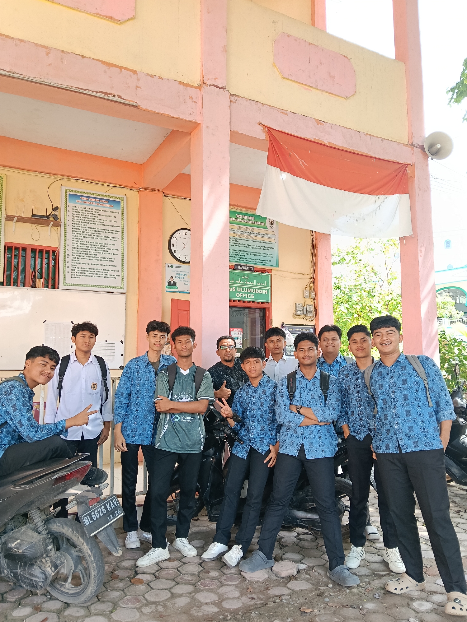 The image depicts a group of ten males posing for a photo in front of a building in Lhokseumawe, Indonesia. Nine of them appear to be students, uniformly dressed in blue patterned shirts and black pants, with varying footwear including sneakers, crocs, and sandals. One student sits on a black Honda motorcycle with the license plate BL 6626 KAV. One male appears to be an adult, wearing a patterned black shirt and glasses, situated amongst the students.
The building features a pink and tan color scheme, with a visible Indonesian flag hanging on the front. Various signs and posters are affixed to the building's exterior, including one titled "TATA TERTIL GURU MTS SWASTA ULUMUDDIN" and another stating "VISI DAN MISI BASAR TSAMANTYAN SWASTA ULUMUDDIN". A clock is also visible on the building. The surroundings include greenery and part of another building in the background. The ground is paved with interlocking hexagonal stones.