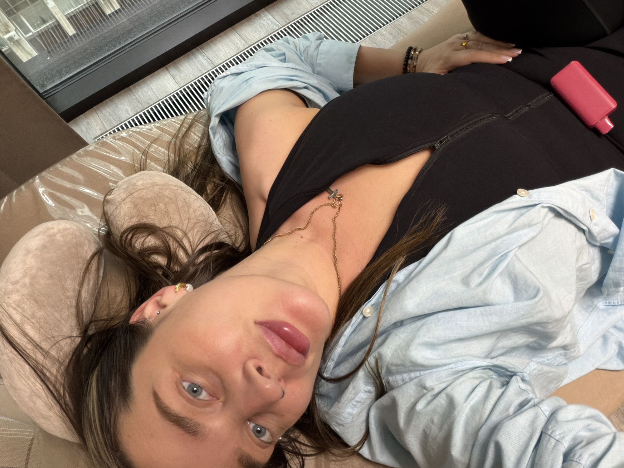 A woman with light skin and light blue eyes is lying on her back on a beige pillow, with brown hair. She wears a black top with a zipper and a light blue button-down shirt. A gold necklace with a cross pendant is visible. She wears earrings and multiple rings. A pink object rests on her black clothing. The background includes a window and a metal grate. The scene is potentially indoors. There is no direct confirmation of the location being Moscow, Russia from the given image.