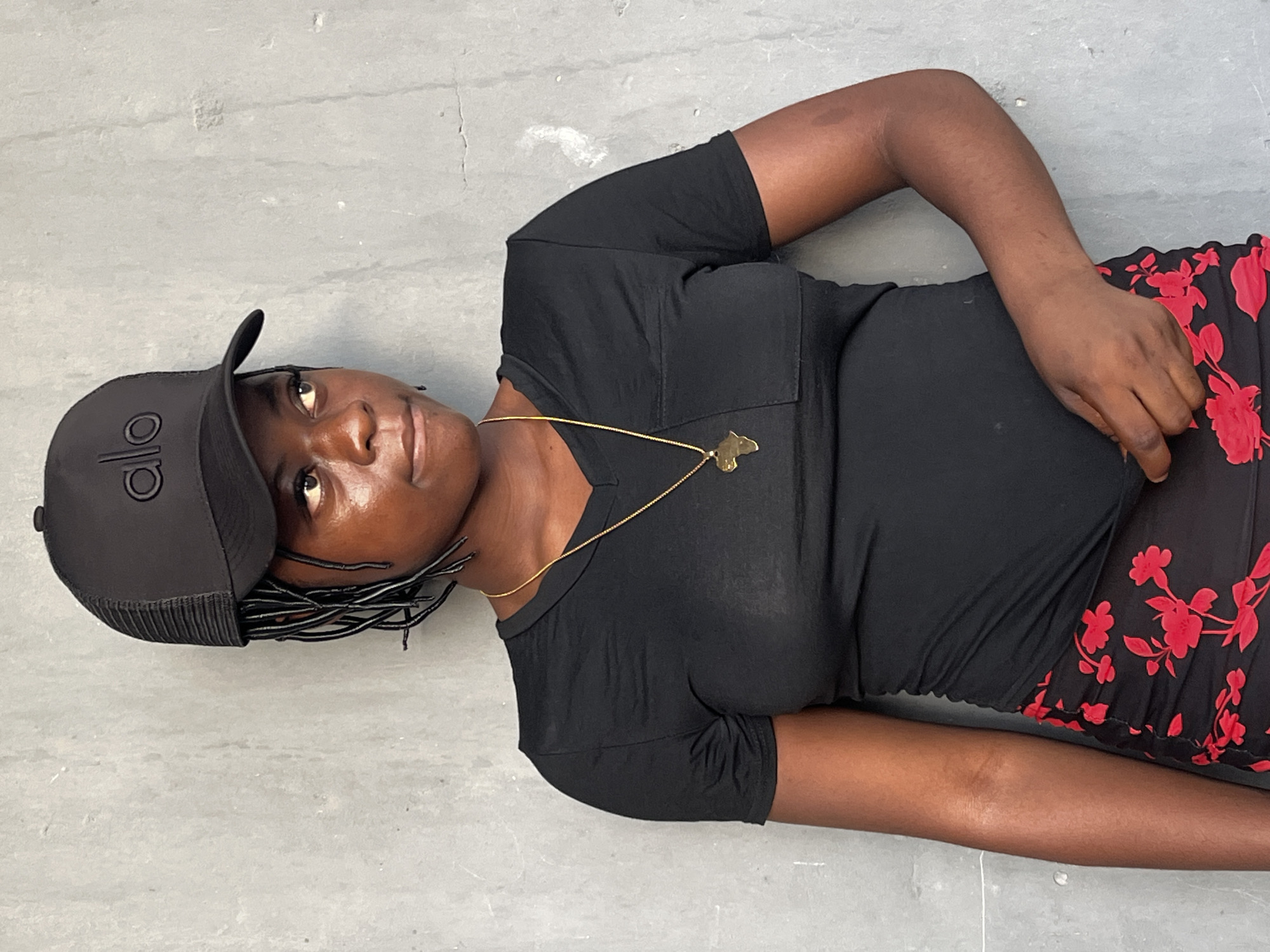 A medium shot captures a dark-skinned woman in an indoor setting. She is lying on a gray surface, possibly a floor. The woman is wearing a black t-shirt, a black cap with a logo, and a floral skirt with red patterns on a black background. She has a gold necklace with a pendant. The scene does not contain enough details to reliably locate the specific location in Badagry, Nigeria.