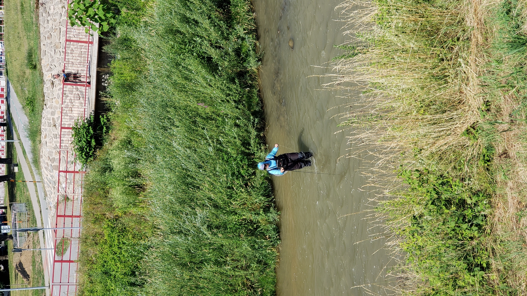 A person is wading in a shallow, murky river, dressed in waders and a light blue jacket, holding a fishing rod. The person is positioned in the middle of the river, with thick green vegetation lining the banks. In the background, a red railing and a stone wall run along the river, with a pedestrian walkway and road above it. Two other individuals are visible on the walkway near the railing. The scene appears to be a natural area within or on the outskirts of a city. The presence of a river, vegetation, and urban infrastructure suggests a location like the Torysa river in Prešov, Slovakia, which has recreational areas and walking paths along its banks.