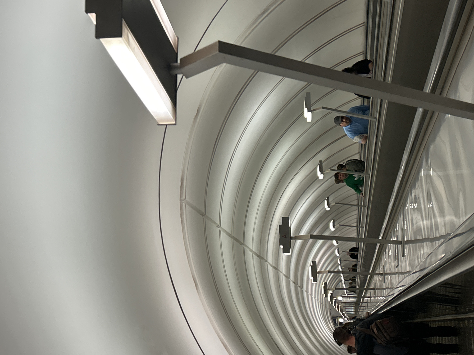 The image captures a tilted view of people ascending or descending a long escalator within a distinctive tunnel-like structure. The perspective is rotated counter-clockwise, making the escalator appear to run diagonally from the bottom right towards the upper left.
The scene is set inside a brightly lit, enclosed space, likely a subway or metro station. The tunnel walls are curved and feature a series of parallel, light-colored ridges or panels that create a ribbed effect, leading to a vanishing point in the distance. Illumination comes from numerous linear lighting fixtures mounted on cantilevered metal brackets that extend from the curved walls, repeating down the length of the escalator. These lights cast a bright, cool glow on the environment.
Several individuals are visible on the escalator steps. In the foreground (bottom right), a person wearing a dark top, headphones, and a cap or hood is seen from behind, carrying a shoulder bag. Further along the escalator, a person in a green long-sleeved shirt and another in a light blue long-sleeved shirt and a dark baseball cap are discernible. Other figures appear further away, becoming less distinct in the distance. The activity depicted is people commuting or traveling on the escalator.
The overall aesthetic is modern and somewhat industrial, with a strong sense of depth created by the repeating architectural elements and lights. No visible text or specific location cues are legible in the image, and the time of day is indeterminate due to the artificial lighting.