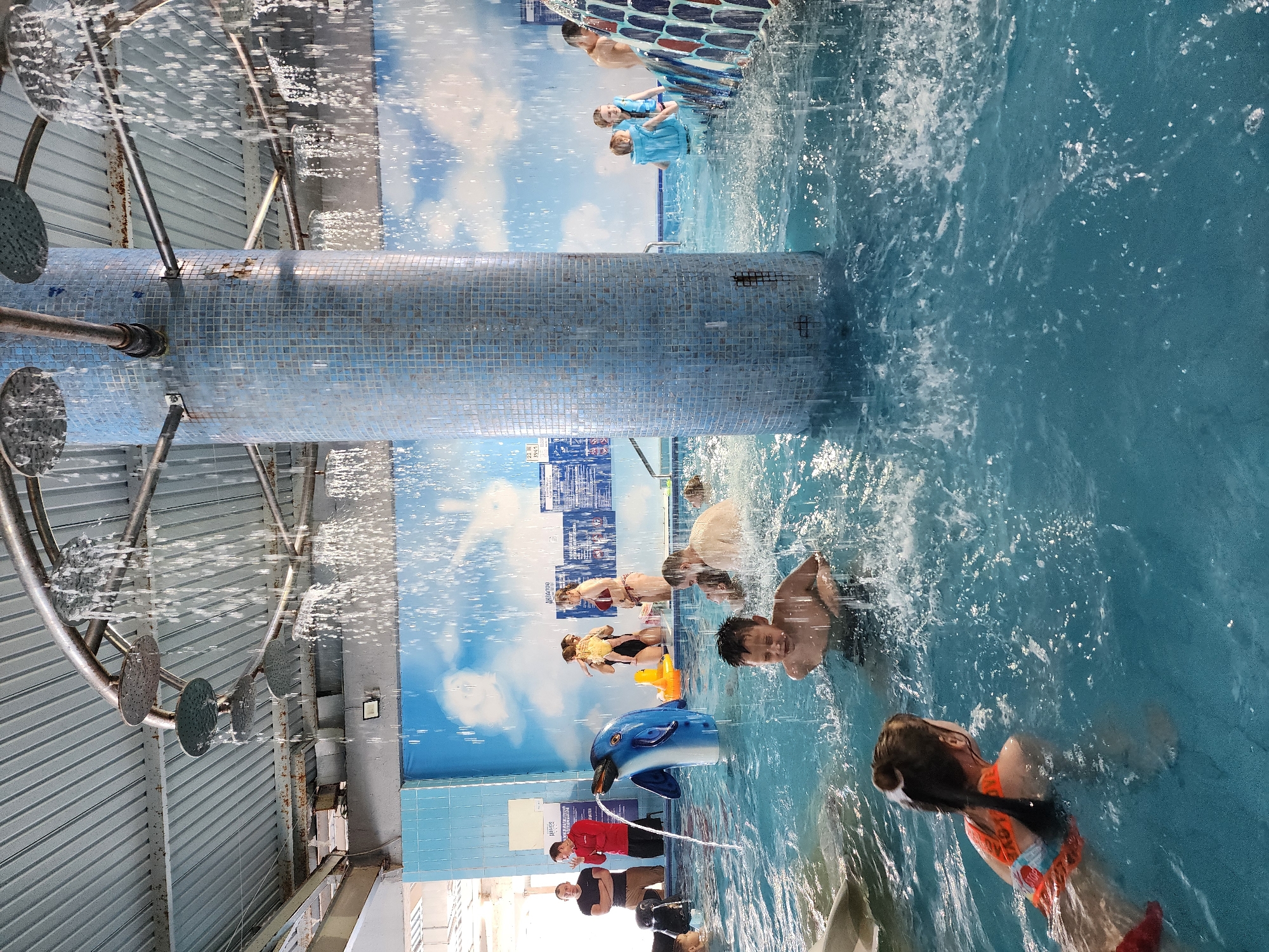 The image shows a vibrant indoor water park, likely in Sochi, Russia, given the Cyrillic characters on some signage. The scene is bustling with activity: several people are swimming and interacting in the bright blue water. Some are clearly enjoying the water features, including an overhead shower structure and a dolphin-shaped water sprayer. The environment is colorful, with a mural of a cloudy sky and city skyline visible in the background, adding to the playful atmosphere. The water park is well-lit, with a mix of natural and artificial light illuminating the scene. A group of adults wearing red shirts near the dolphin feature suggests the presence of park staff or lifeguards.