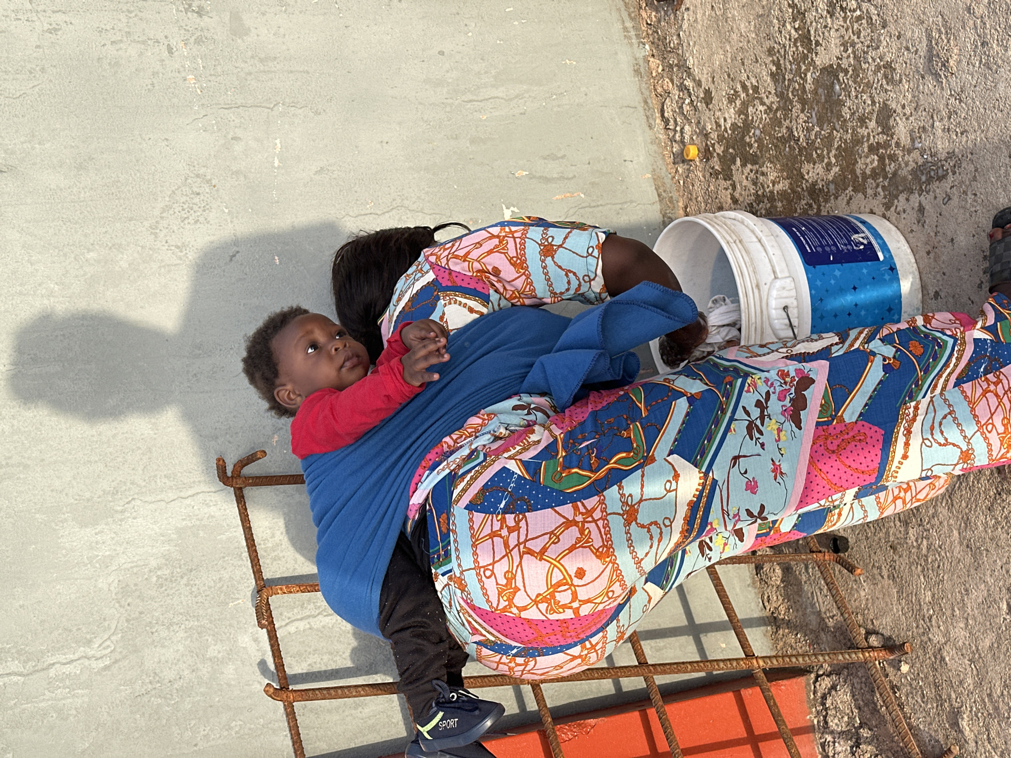 The image, taken in Gura Top, Nigeria, depicts a woman and a baby in an outdoor, sunlit setting. Strong shadows suggest it is daytime.
The woman, whose face is not visible, is wearing a vibrant, multi-colored patterned top and matching pants, adorned with chain and floral designs in blue, pink, orange, and white. Her dark hair is pulled back, and she wears black sandals. She appears to be bending or leaning over, possibly attending to an item on the ground.
She is carrying a baby or young child in a blue fabric wrap or carrier. The baby, with dark skin and short dark hair, is wearing a red long-sleeved shirt and black pants, and a black sneaker with a yellow stripe and the word "SPORT" on its side. The baby is facing away from the woman's body, looking upwards with a neutral expression.
The ground is a light gray concrete or paved surface, showing some wear and rougher, darker patches. A rusty metal rebar grate or frame is prominent in the foreground. To the right, a white plastic bucket with a blue label lies on its side. The label features a star pattern and the partially visible brand name "DULUX". A small yellow object is also visible on the ground nearby. The background consists of a concrete or stucco wall, exhibiting some cracks and discoloration.