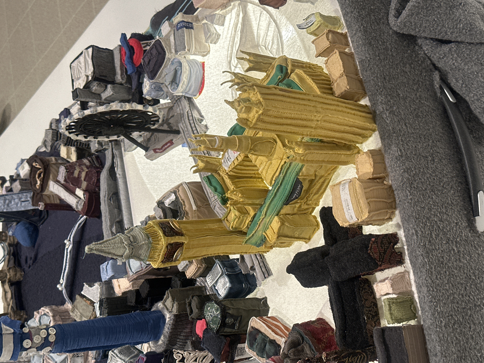 The image depicts a mixed media artwork on display, possibly within a gallery or museum setting in London, United Kingdom. The artwork is composed of various fabric scraps and textile elements arranged in a three-dimensional, collage-like fashion. Dominant colors include shades of brown, green, blue, grey, and tan, with a variety of textures and patterns evident in the textiles used.
The artwork features recognizable shapes, including geometric forms and possibly representational elements such as clothing and architectural fragments. A black, circular object with a radial pattern is visible, alongside a section of a grey wool garment. The artwork appears to be mounted on a white surface, likely a wall. The composition is dense and complex, with individual textile pieces layered and overlapping to create an overall textured and multi-faceted effect.