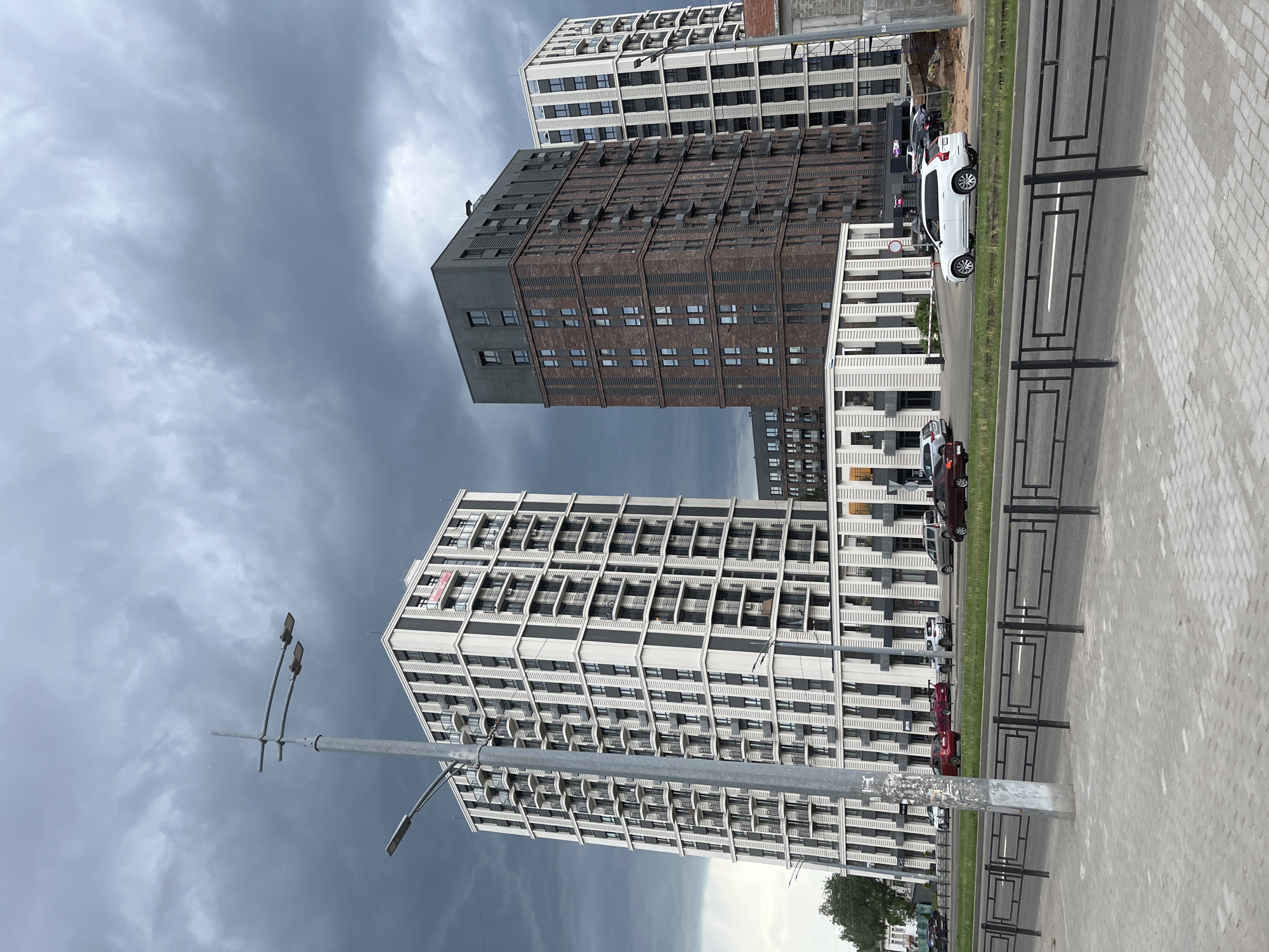 The image shows three high-rise buildings in Izhevsk, Russia. The buildings are predominantly white and gray with some brown elements. A row of parked cars are visible beside the buildings, separated by a strip of grass from a walkway. A metal street light pole stands in the foreground, and a dark, cloudy sky forms the backdrop.