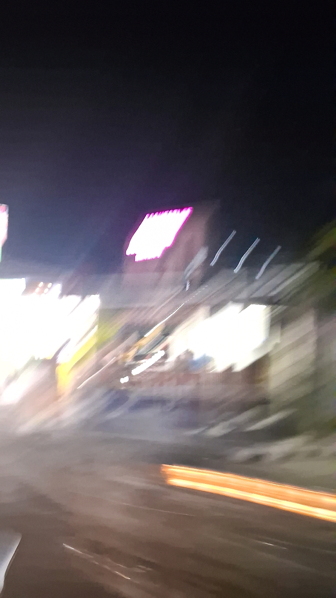 The image is a blurred, nighttime shot of what appears to be a street scene in Berbah, Indonesia. The motion blur suggests the photograph was taken from a moving vehicle. In the upper portion of the image, there are bright, rectangular neon or LED signs, one of which is pink and appears to say "SUMBER". Other blurry, bright lights and shapes indicate buildings or structures lining the street. The ground is dark and appears to be asphalt, with some faint white lines suggesting road markings. A distinct streak of orange light is visible horizontally across the lower right quadrant, likely from vehicle headlights or taillights. The overall impression is of a busy, illuminated urban environment at night, with the motion blur contributing to a sense of speed and dynamism.