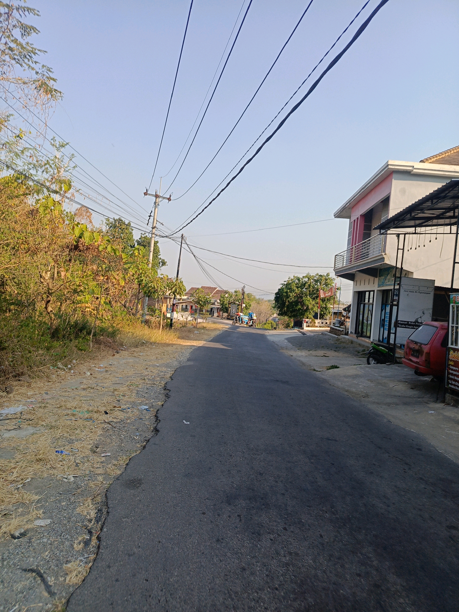 The image depicts a street scene in Ringinagung, Indonesia, under a clear sky. A paved road runs through the center of the frame, bordered on the left by a dry, overgrown embankment with sparse vegetation and scattered debris. On the right side of the road, there is a multi-story building with a modern facade, featuring a balcony on the upper floor and glass storefronts on the ground level. A dark-colored car and a green motorcycle are parked in front of the building. Further down the road, several houses and trees are visible, along with utility poles and overhead power lines crisscrossing the sky. A few people are seen on motorcycles in the distance, suggesting ongoing activity in the area. The overall impression is of a rural or semi-urban residential and commercial area during daylight.