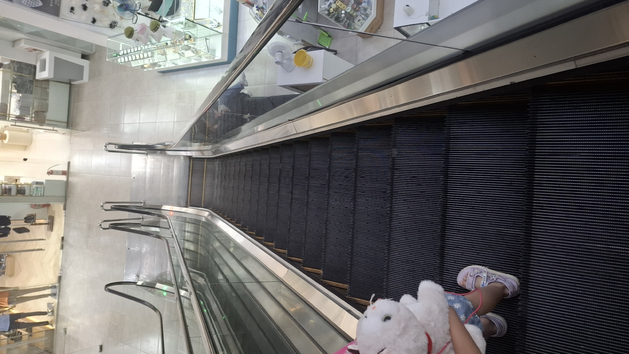 The image shows an overhead view of an escalator in a retail setting, possibly a mall or department store in Shymkent, Kazakhstan. The escalator is moving downwards, indicated by the horizontal steps. A young child, facing away from the camera, is riding the escalator. They are wearing blue denim shorts with white stars and light purple sandals. The child is holding a white stuffed animal. The escalator has a metal handrail and glass panels on the sides.
To the left of the escalator, a bright, tiled floor is visible, reflecting the overhead lights. In the background, displays of merchandise can be seen through glass cases. One display features jewelry or accessories. Another area shows shelves with various items. A fire extinguisher is mounted on a white wall. The overall lighting suggests an indoor environment, likely during daylight hours. The scene conveys a sense of everyday activity within a commercial space. No visible text is present in the image.