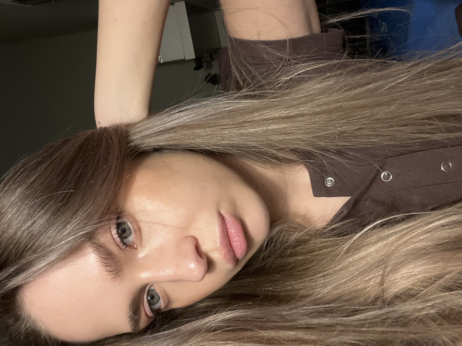 The image features a close-up portrait of a young female subject. She has long, straight to wavy light brown hair with blonde highlights, parted on her left side, which falls across her shoulders and chest, extending through the lower frame. Her eyes are blue-green, and she has naturally full, light brown eyebrows. Her facial features include a straight nose and full lips with a soft pink tint. Makeup application is subtle, including light pink eyeshadow and a luminous finish on her skin, particularly on her left cheekbone. She is wearing a dark brown, ribbed, short-sleeved top with a placket secured by three visible silver snap buttons. Her left arm is raised, with her hand partially visible near the back of her head, appearing to touch or adjust her hair. The subject is looking directly into the camera with a neutral expression. The background is dark and out of focus, indicating an indoor setting. Indistinct light-colored furniture or shelving is faintly visible in the upper right portion of the background. The specific location within Saint Petersburg, Russia, cannot be determined from the visual information present in the image.