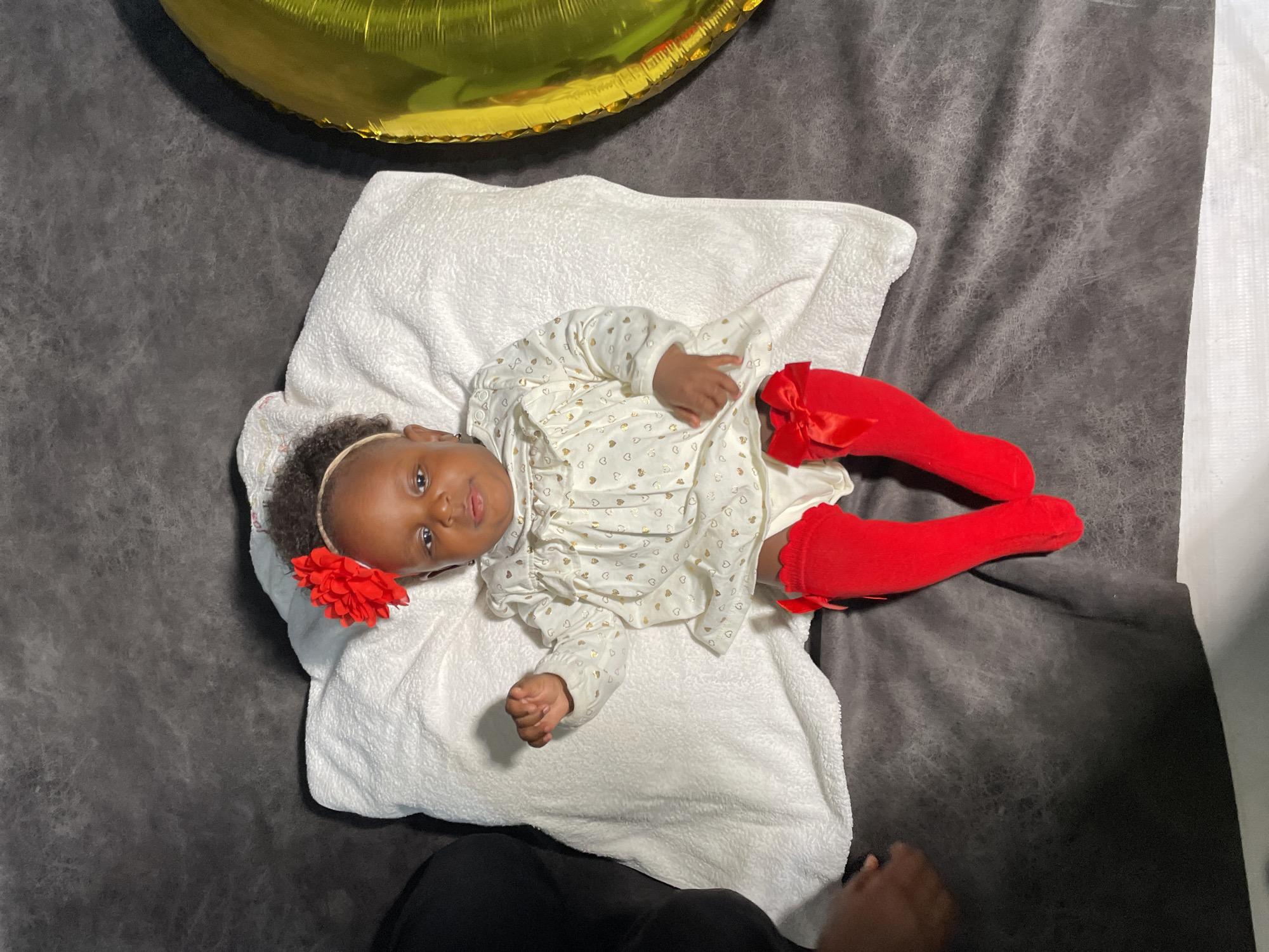 A baby lies on a white pillow on a grey surface, presumably a blanket or rug. The baby is wearing a white dress with gold heart patterns and red leggings with bows on the sides. A red flower is clipped to the baby's hair. A partially visible gold balloon is in the upper left corner. The location is not discernible from the image, and there is no indication it is in Barakin Akawo, Nigeria.