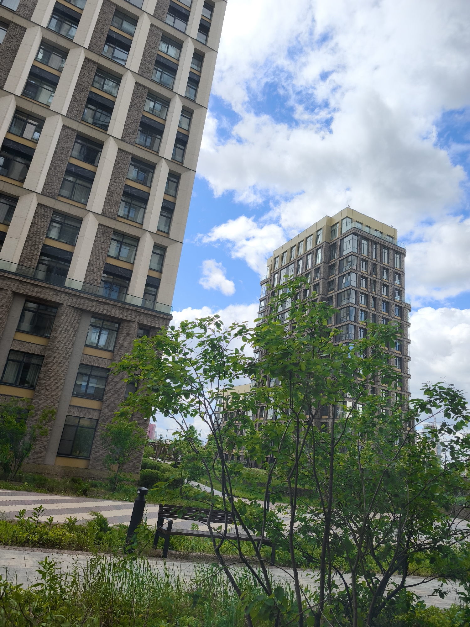 The image depicts an outdoor scene in an urban setting, likely in Moscow, Russia. Two modern high-rise buildings with tan-colored facades and dark windows dominate the backdrop, set against a partly cloudy blue sky. The building on the left is angled in the frame, while the one on the right appears more upright. In the foreground, a green, tree-filled park area is visible, partially obscuring a wooden park bench. The ground is covered with green grass and small plants. A section of paved walkway is visible in the lower left corner of the frame.
