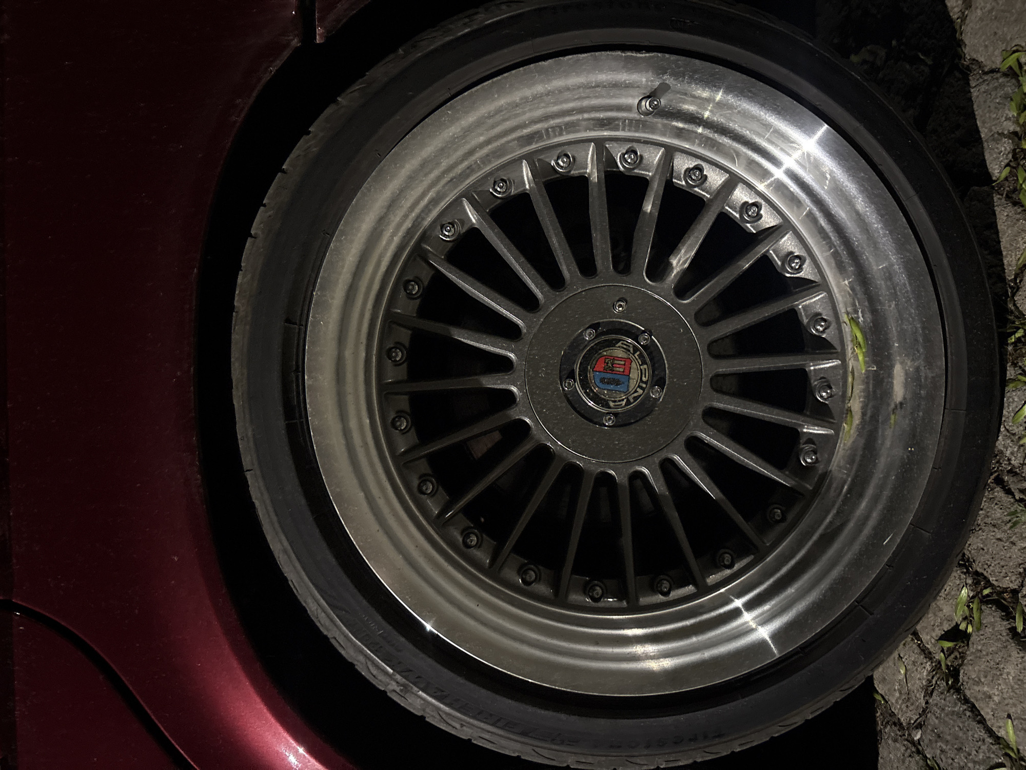 A close-up image shows a wheel and tire mounted on a maroon-colored vehicle. The wheel features a multi-spoke design with a prominent center cap displaying an emblem. The tire is black and appears to be new or in good condition. The vehicle is parked on a surface that appears to be a cracked sidewalk or concrete with some vegetation. The location is not discernible from the image and there is no indication of being in Sofia, Bulgaria. There are no visible people or indications of interactions in the frame. The image focuses primarily on the aesthetic and mechanical details of the vehicle wheel.