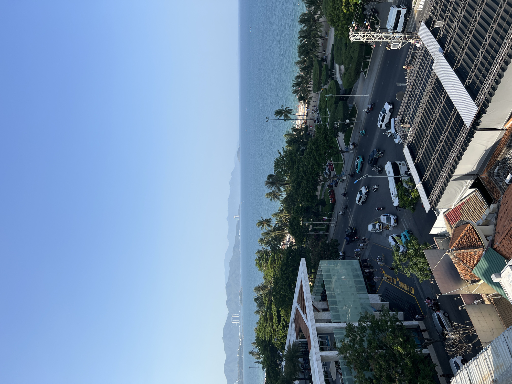 This aerial view captures a coastal city scene in Nha Trang, Vietnam, on a clear, bright day. The foreground is dominated by the rooftops of buildings, some with corrugated metal and others with tiled roofs. A multi-story modern building with a white facade and glass panels is visible on the left. Adjacent to it is a street bustling with traffic, including cars, motorcycles, and a bus. People are seen walking along the sidewalk and crossing the street.
Beyond the street, a lush green expanse of trees, including palm trees, separates the city from the beach. The beach itself is populated with people enjoying the sun and sand. The turquoise waters of the ocean stretch to the horizon, where a hazy mountain range forms the backdrop. A few small boats are scattered across the sea. The overall impression is one of a vibrant coastal city with a clear blue sky overhead.