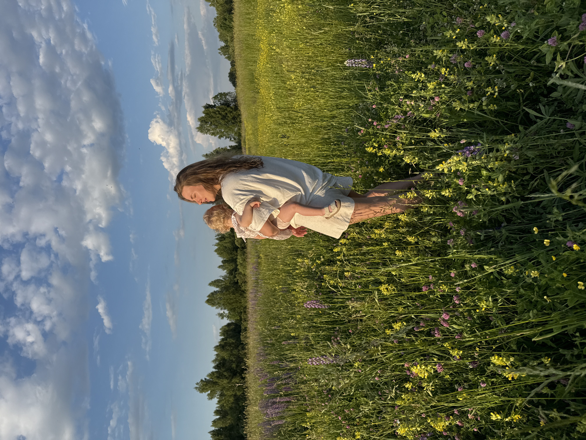 In a sunlit meadow in Koloksha, Russia, a woman with long, wavy brown hair holds a young child. The woman, wearing a loose, off-white outfit and sandals, gazes into the distance, her eyes closed. The child, in a light-colored dress, is held in her arms. The meadow is filled with tall green grass, interspersed with yellow and purple wildflowers. In the background, a line of trees borders the field, and a bright blue sky with scattered white clouds stretches above. The scene captures a moment of quiet connection between mother and child in a natural, rural setting.